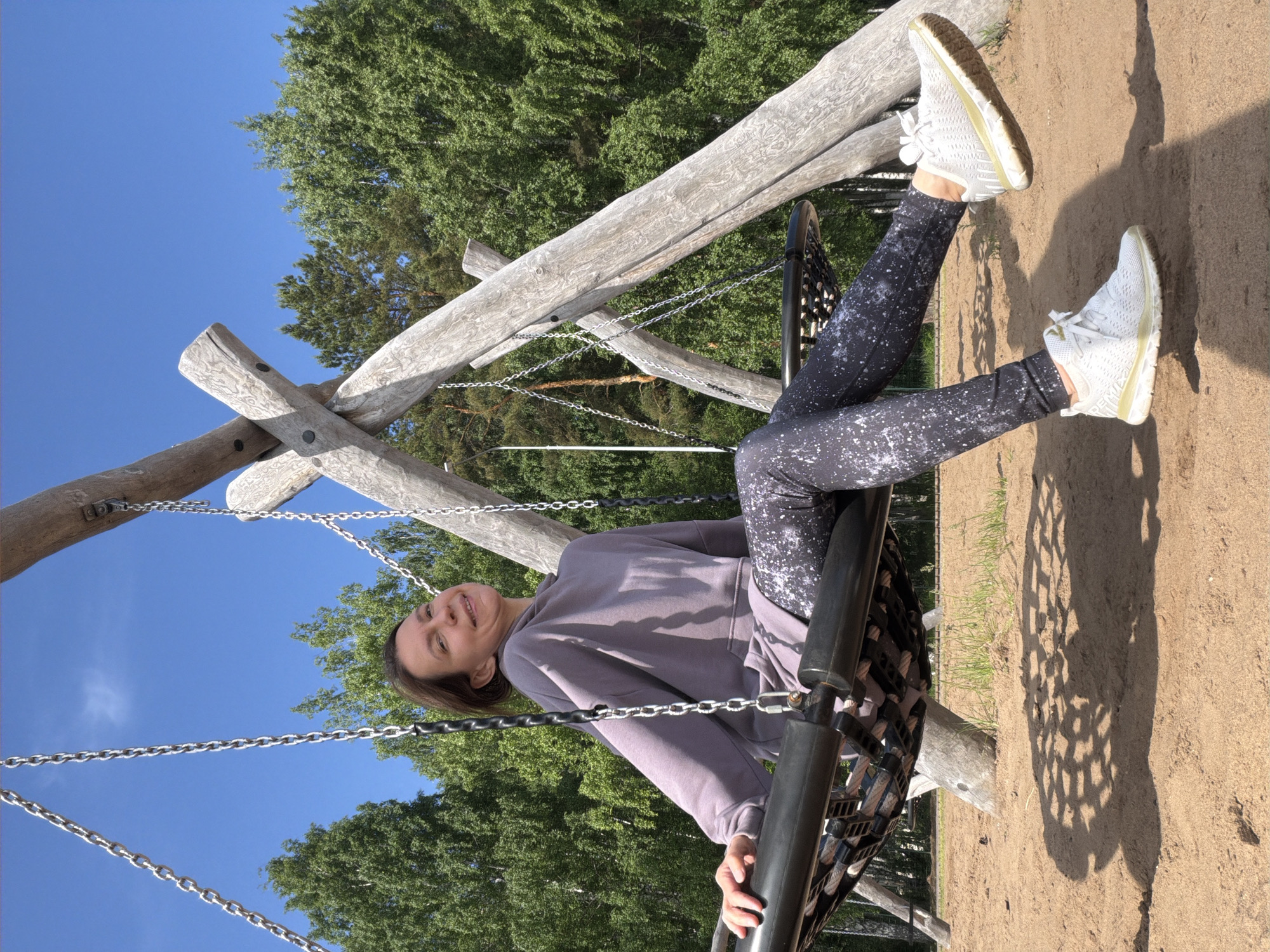 An outdoor, eye-level shot depicts a woman swinging on a large wooden swing set under a clear blue sky in Chaikovsky, Russia. The image is oriented with the woman lying horizontally, head to the left, feet to the right, as if seen from beneath the swing.
The central subject is a young adult woman with light brown hair, smiling brightly and looking slightly upwards and to her left. She is wearing a light purple-grey hooded sweatshirt and black leggings with white speckled patterns. Her legs are extended upwards, with her feet clad in white athletic sneakers that have light yellowish soles. She appears relaxed and happy, holding onto the swing's side support with one hand.
The swing set is constructed from robust, light-colored wooden logs, featuring a natural, rustic appearance with visible grain. The woman is seated in a large, round, basket-style swing seat made of woven material, suspended by sturdy metal chains from the wooden frame.
The background features lush green trees with dense foliage, indicating a natural or park-like setting. The ground beneath the swing is sandy or composed of fine dirt, with patches of sparse green grass visible. Bright sunlight casts distinct shadows on the ground, including a patterned shadow from the swing's base, suggesting it is daytime, likely mid-morning to late afternoon, with clear and sunny weather. The overall atmosphere conveys joy and outdoor leisure. No visible text is present in the image.