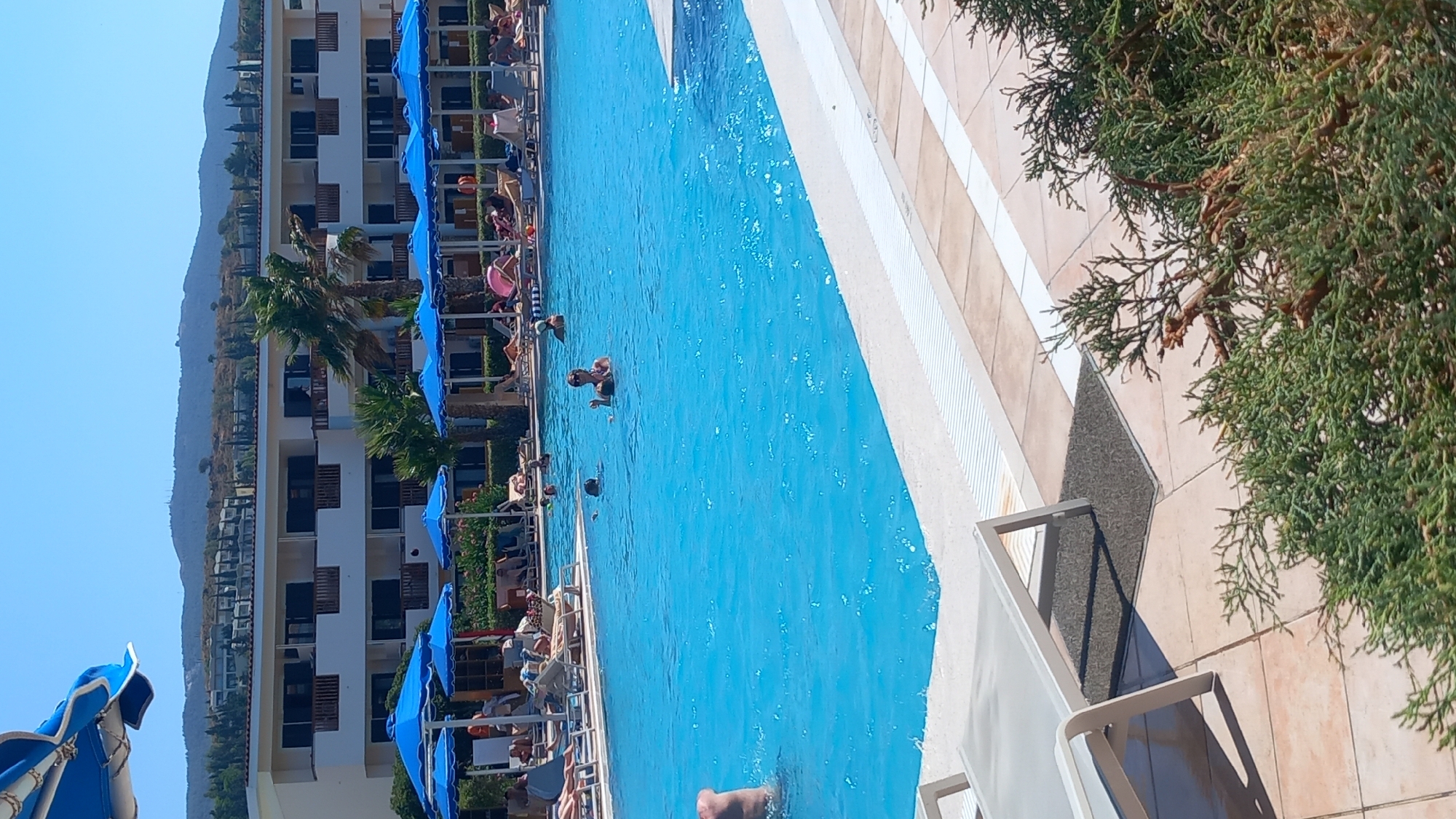 This image shows a resort swimming pool in Kos, Greece. The pool is a bright blue, and the surrounding area is paved with light brown tiles. Several blue umbrellas are set up around the pool, providing shade for lounge chairs. In the background, there is a multi-story hotel building with balconies and windows. The hotel is surrounded by lush green trees and bushes, with a mountainous landscape visible in the distance under a clear blue sky. There are several people in the pool, some swimming and others standing in the shallow end. One woman in the foreground is standing in the water, facing the camera with sunglasses on. Another person's leg is visible emerging from the water on the left side of the frame. Lounge chairs are visible on the paved area surrounding the pool. In the foreground, on the right, a green bush with branches is partially obscuring the view.
