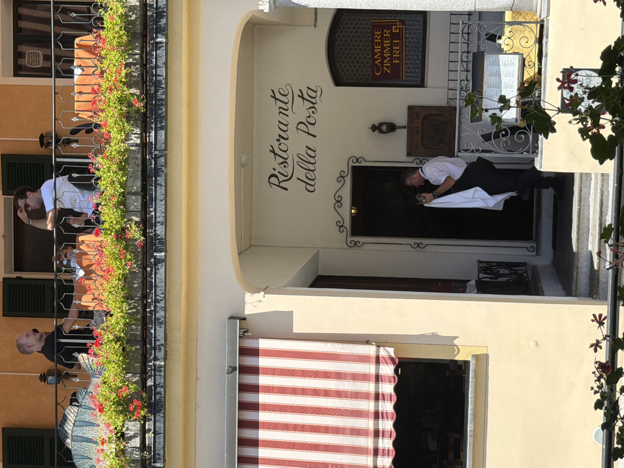 The image depicts the exterior of "Ristorante della Posta," a restaurant located in Lugano, Switzerland, as indicated by the signage. Above the restaurant's entrance, a balcony is visible with people on it and flower boxes filled with red flowers and greenery. The restaurant itself features an awning with red and white stripes above a window. A man in a white shirt and black pants, presumably a waiter, is seen exiting the building carrying a white cloth. A sign on the wall reads "CAMERE ZIMMER FREI," suggesting available rooms. The overall scene suggests a sunny day, possibly in a European town.