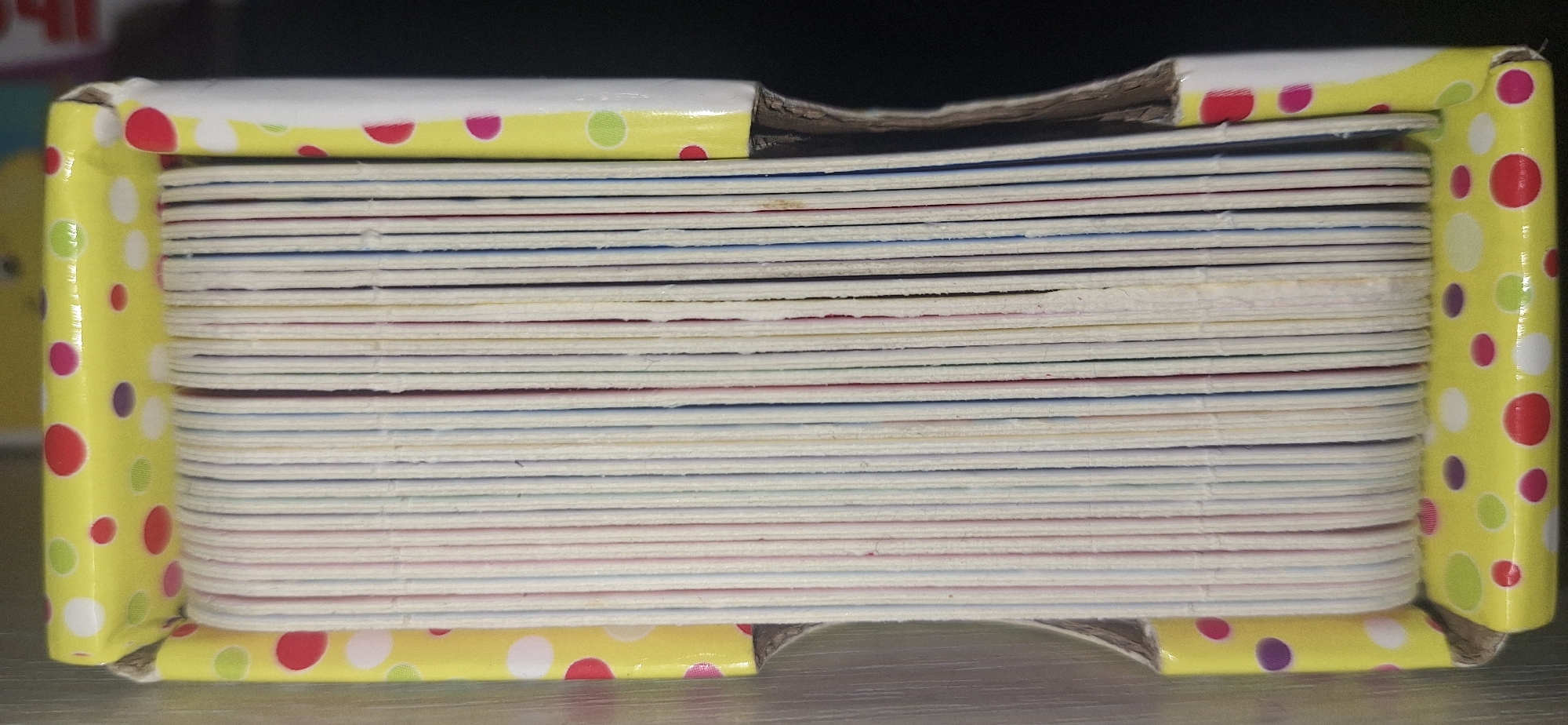 The image shows a close-up, side view of a thick stack of cards or thin books contained within a decorative box. The box is yellow with a pattern of colorful polka dots in red, pink, green, and purple. The edges of the box are visible, wrapping around the stack of cards. The stack of cards is tightly packed, with the edges of each individual card showing distinct lines. Some of the cards appear to have colored edges, with thin stripes of red, blue, and possibly other colors visible between the white cardstock. The cardboard interior of the box is slightly exposed in some areas, particularly at the top where it seems a bit damaged. The lighting is even, suggesting an indoor setting, and there are no discernible time-of-day or weather cues. There is no visible text in the image, nor are there any people, animals, or specific activities depicted. The focus is solely on the object itself, a stack of cards within its packaging. The setting is likely a shelf or table in a room, with a dark background that contrasts with the bright yellow of the box.