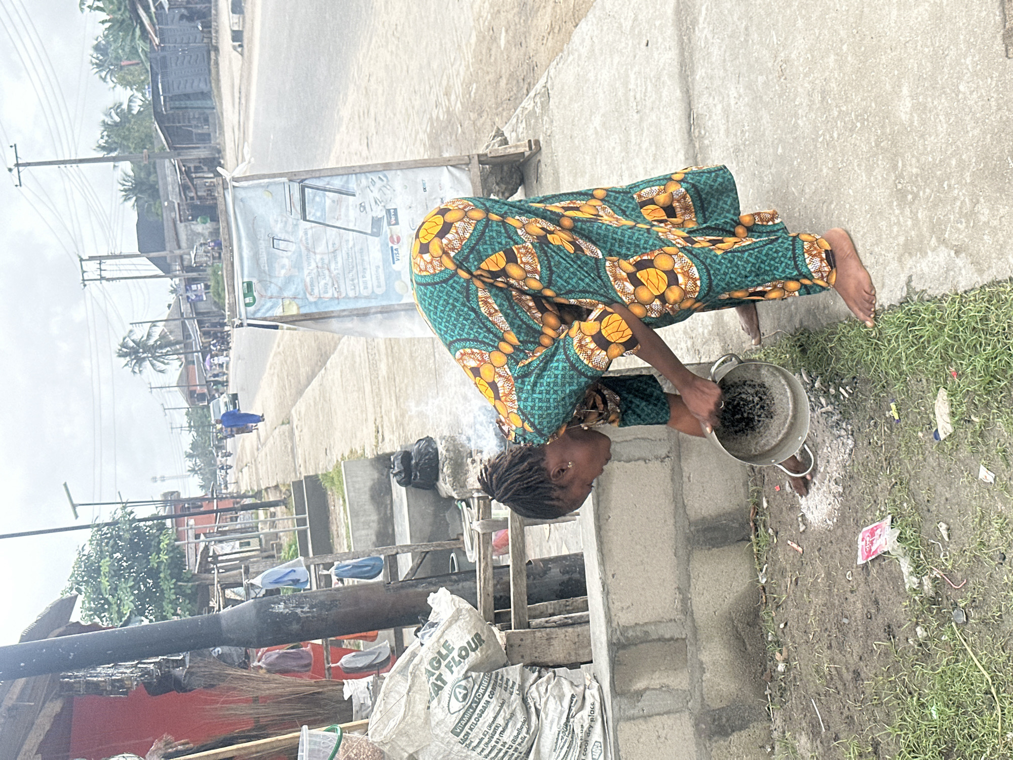 In Badagry, Nigeria, a woman in a vibrant, patterned green and orange dress is bending over a low concrete wall. She is pouring ash from a metal pot onto the ground. The pot appears to have some ash still clinging to its interior. Her hair is styled in dreadlocks. In the background, a dusty road stretches out, lined with buildings and scattered vegetation. A few people are visible in the distance, and a vehicle is driving on the road. To the left of the woman, a large bag of "Eagle" brand flour is stacked against a wooden structure. The scene appears to be a street market or an outdoor vendor stall.
