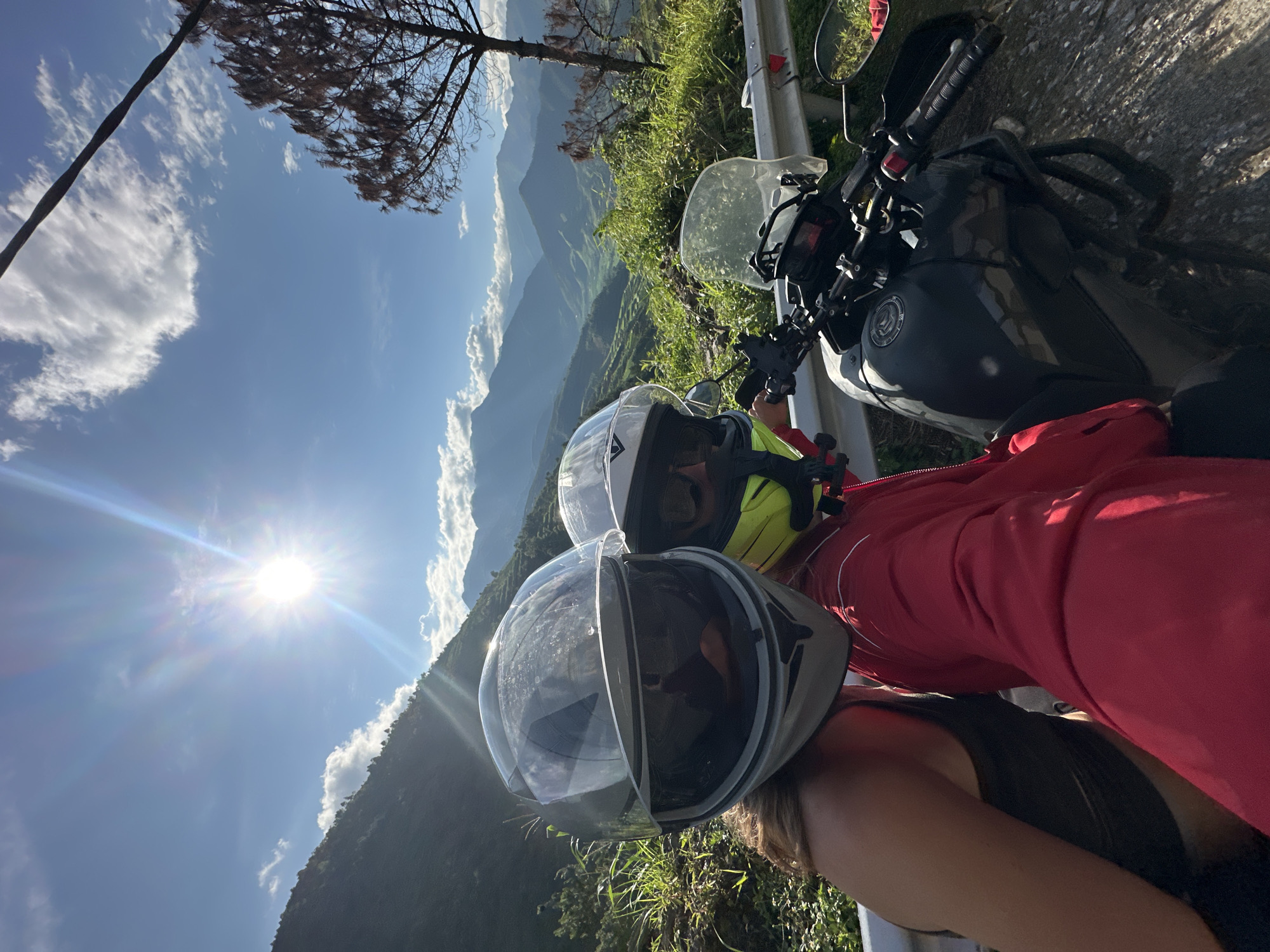 Two individuals in motorcycle helmets are depicted in a selfie-style shot, taken on a sunny day in a mountainous region identified as Tú Lệ, Vietnam.
The subject on the left wears a light gray full-face motorcycle helmet with its clear visor partially raised, revealing dark sunglasses underneath. Their left shoulder and a dark tank top are visible. The subject on the right, partially obscured by the left individual and positioned slightly behind, wears a white and yellow full-face helmet, also with dark sunglasses visible. They are attired in a red long-sleeved jacket.
A dark gray or black adventure-style motorcycle is parked beside them on a paved road. Visible components include its handlebars, a clear front windshield, fuel tank, and engine crash bars. The motorcycle is positioned next to a silver metal guardrail.
The environment features a clear blue sky with scattered white clouds. A prominent sunburst emanates from the upper-left sky, casting strong light across the scene. The background consists of multiple layers of green, tree-covered mountains, receding into a hazy blue distance. Green terraced fields are discernible in the valleys between the distant mountains. Lush green vegetation and shrubs line the immediate roadside embankment. On the far right, a solitary, defoliated tree with dark, intricate branches stands against the sky.