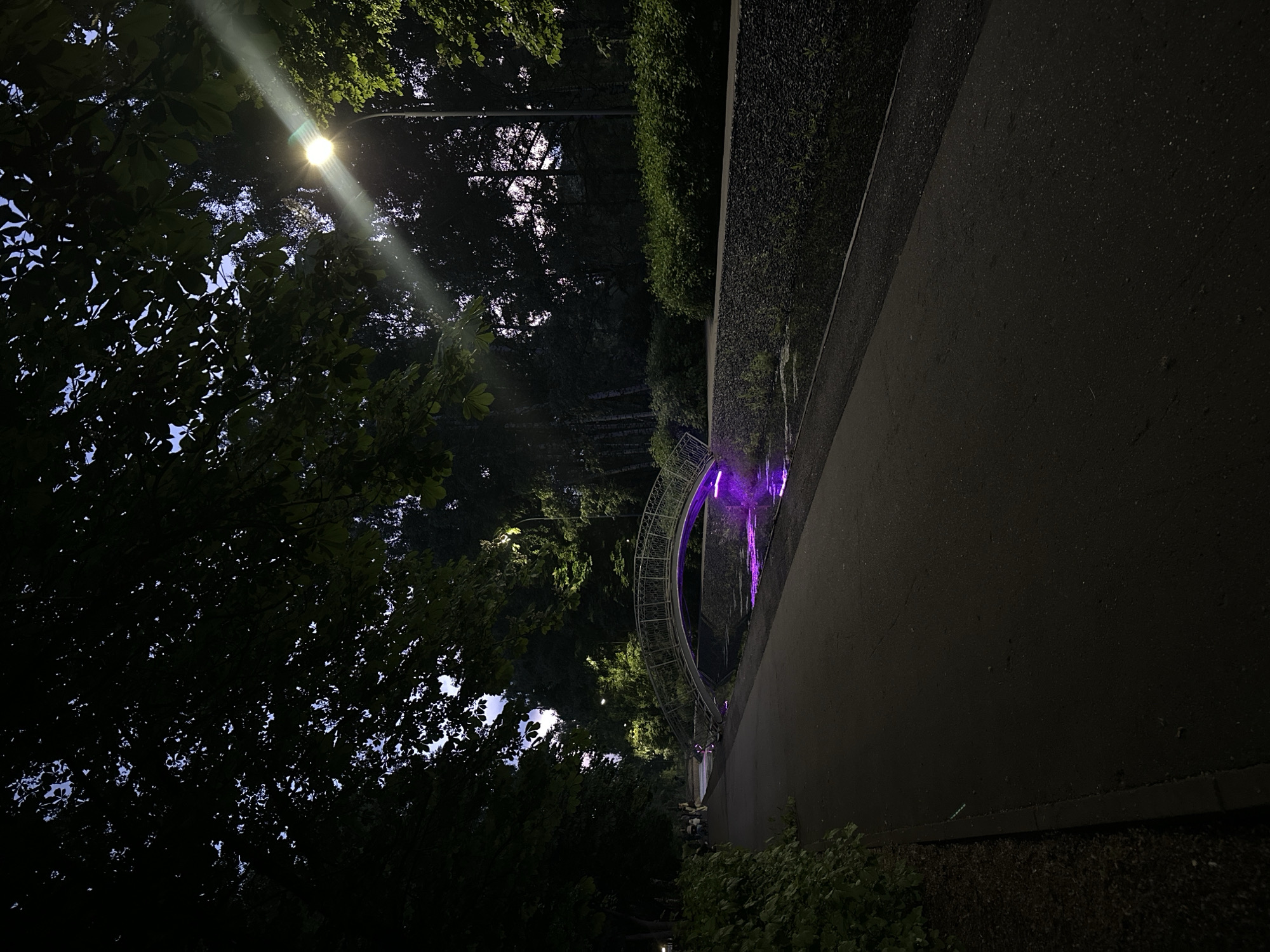 This media file captures a dark, nighttime scene in what appears to be a park or landscaped urban area, identified as Kazan, Russia. The image is dominated by the silhouettes of dense trees and foliage, with glimpses of a lighter sky visible through the leaves.
A bright street lamp is prominently positioned in the upper left, emitting a strong, diagonal beam of light that cuts across the frame and illuminates portions of the tree canopy. Below and to the right of the trees, a dark, paved path or road extends from the foreground into the midground.
A notable feature is a curved, ornate metal structure, possibly a decorative archway or bridge, situated in the midground on the right side, above the path. This structure is brightly illuminated with vibrant purple lights, creating a striking contrast against the dark surroundings. The purple light appears to reflect faintly on the path below, suggesting a wet surface, possibly from recent rain or dew. To the right of the path, a dark wall or elevated platform, topped with a neatly trimmed hedge, adds to the structured landscape.
The scene conveys a tranquil and somewhat enchanting atmosphere, characteristic of a well-maintained public space at night. No people or visible text are present.