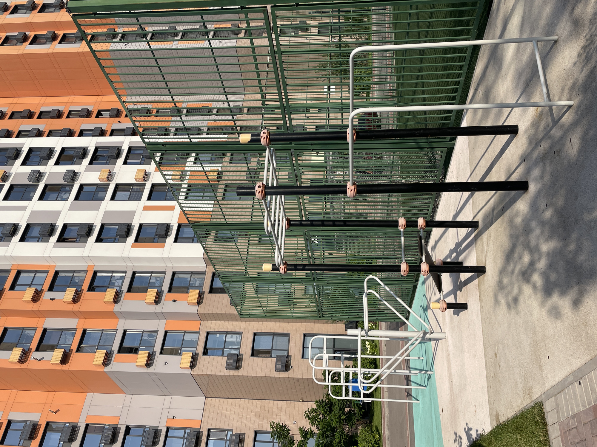 The image depicts an outdoor exercise area with various fitness equipment, including parallel bars and pull-up bars. The equipment is predominantly black with some white components, and the ground is a light-colored concrete. Behind the exercise area is a tall, modern apartment building with a distinctive orange and grey facade, featuring numerous windows and air conditioning units. A green metal fence encloses part of the area, separating it from a sports field with artificial turf. The scene is set during daylight, with shadows cast by the equipment and surrounding structures. The location is identified as Almaty, Kazakhstan.