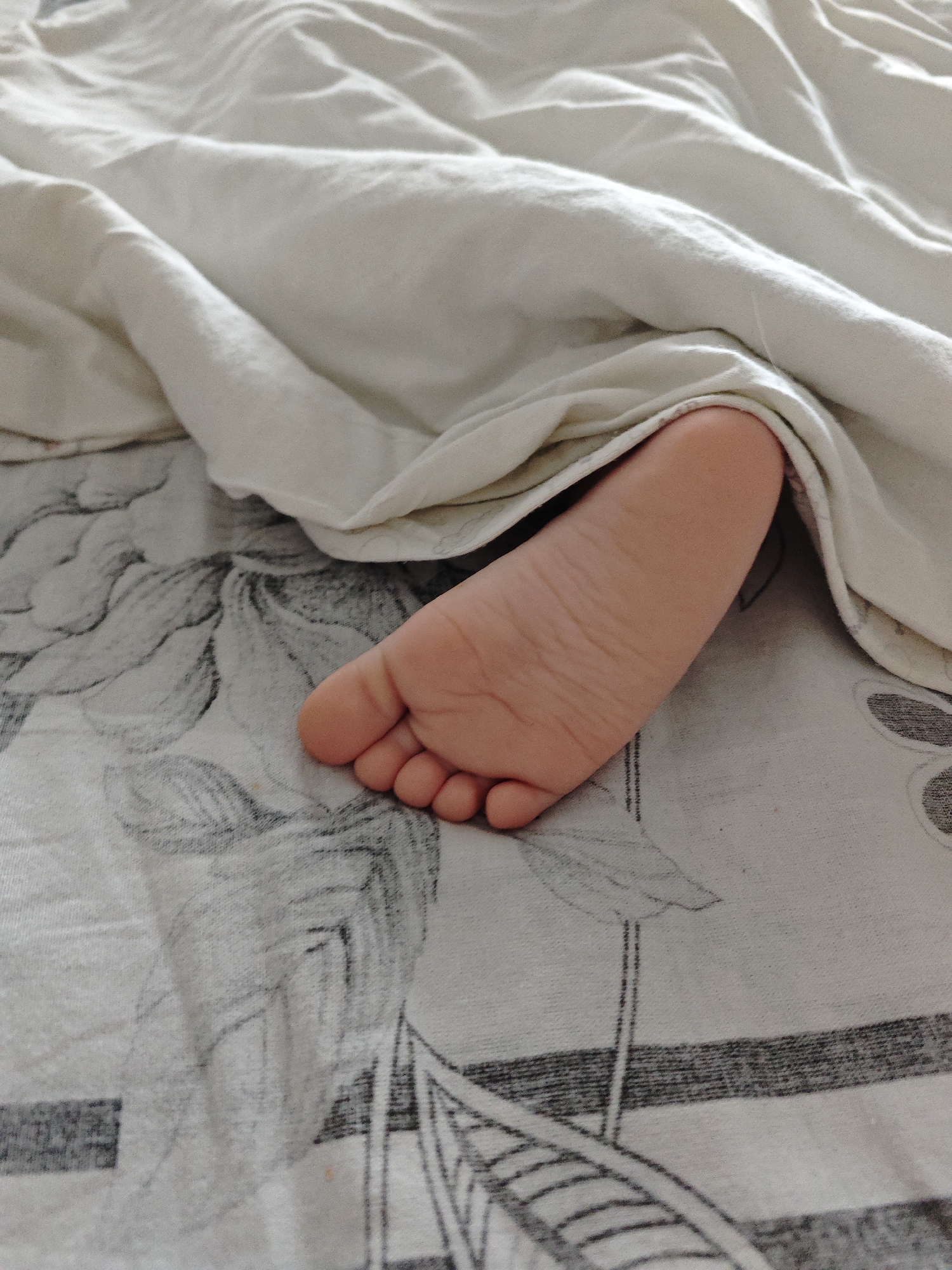 A baby's foot is visible, partially covered by a white sheet on top of a patterned bedsheet. The location is not discernible from the provided image.