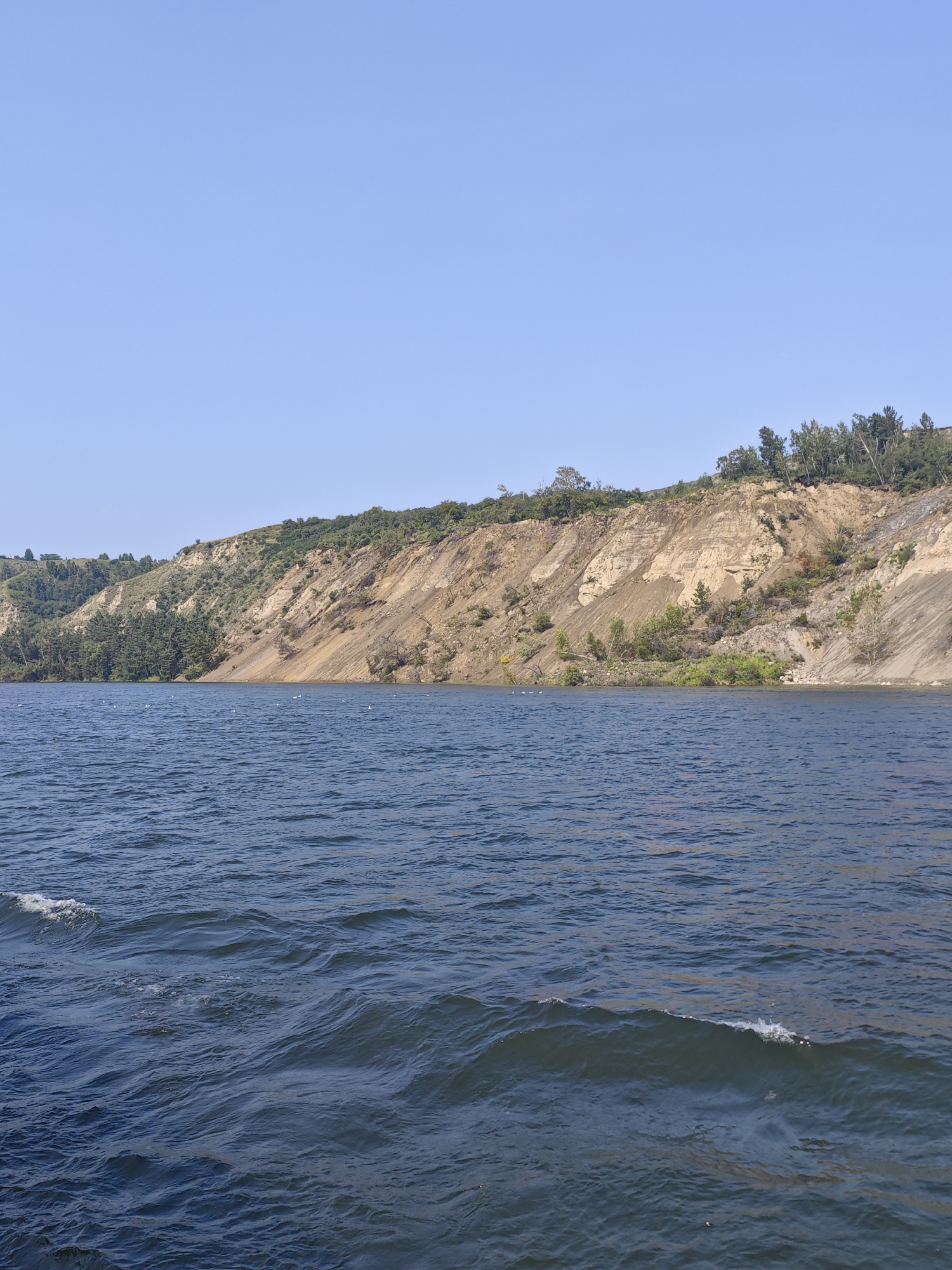 A body of water, likely a lake or reservoir, is in the foreground with moderate waves. In the background, a steep, eroded cliff face rises from the water's edge. The cliff exhibits distinct layers of soil and rock, with patches of vegetation growing on its slopes and crest. A clear blue sky dominates the upper portion of the image. The scene appears to be a natural landscape with no visible man-made structures or human activity. The location is identified as Yermolayevo, Russia, suggesting this is a natural landmark or recreational area within or near the city.
