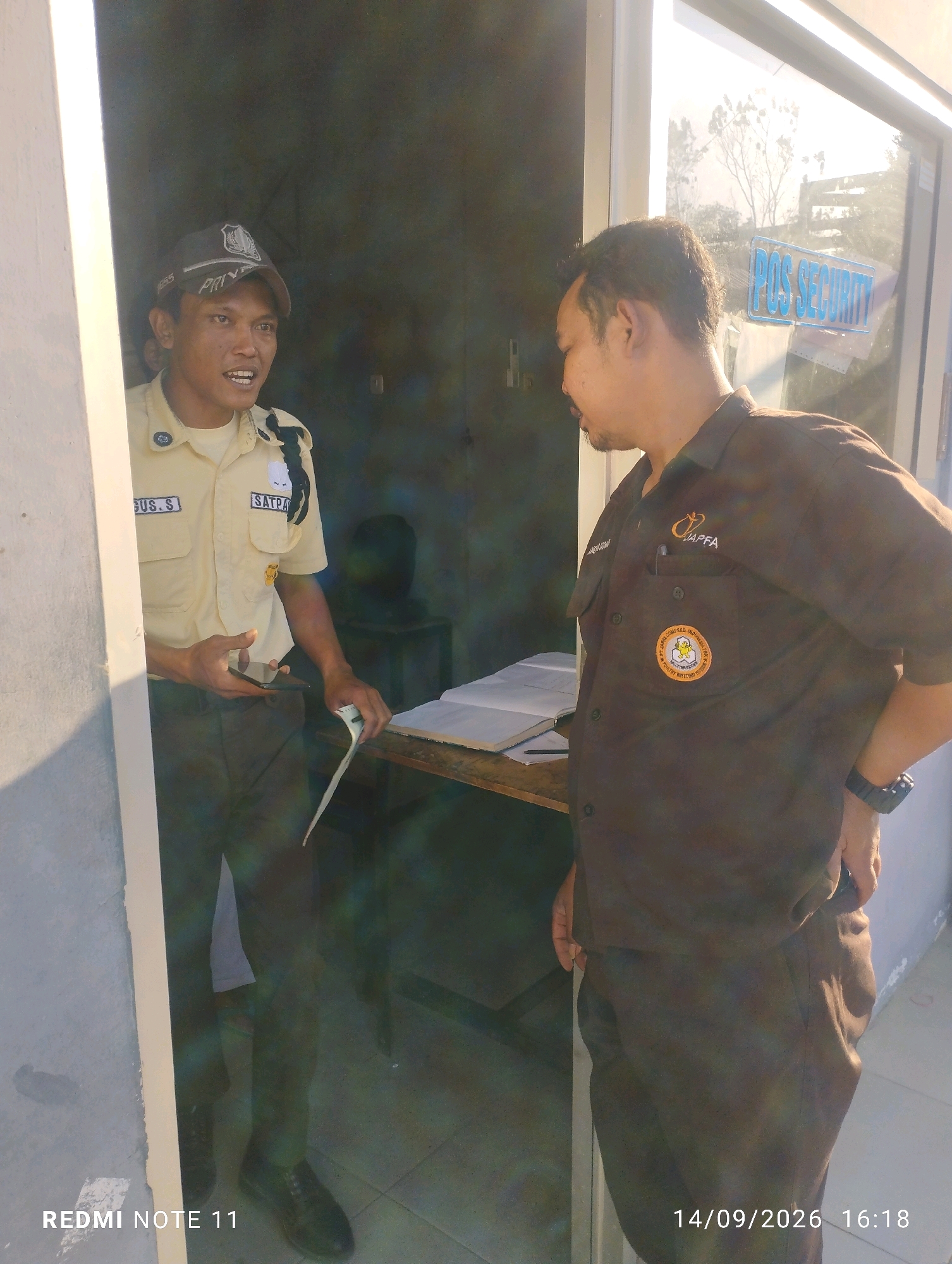 In Bero, Indonesia, two men are interacting in what appears to be an outdoor setting near a building with a glass door. The man on the left is wearing a light-colored uniform with a dark vest and a cap. He has a name tag that reads "GUS. S" and another that says "SATPAM". He is holding a mobile phone in his left hand and a piece of paper in his right hand. He is looking towards the man on the right and appears to be speaking.
The man on the right is wearing a dark brown collared shirt with a logo on the left chest that reads "PAPFA". He also has a circular patch on his left breast with an image of a chicken and text around it. He is facing the man on the left and appears to be listening. His left hand is on his hip, and his right hand is near his pocket.
Behind the men, there is a table with some papers and a pen on it. Through the glass door, trees and a sign that says "POS SECURITY" are visible. The lighting suggests it is daytime, with sunlight illuminating the scene. The overall environment appears to be functional, possibly an entrance or security post.