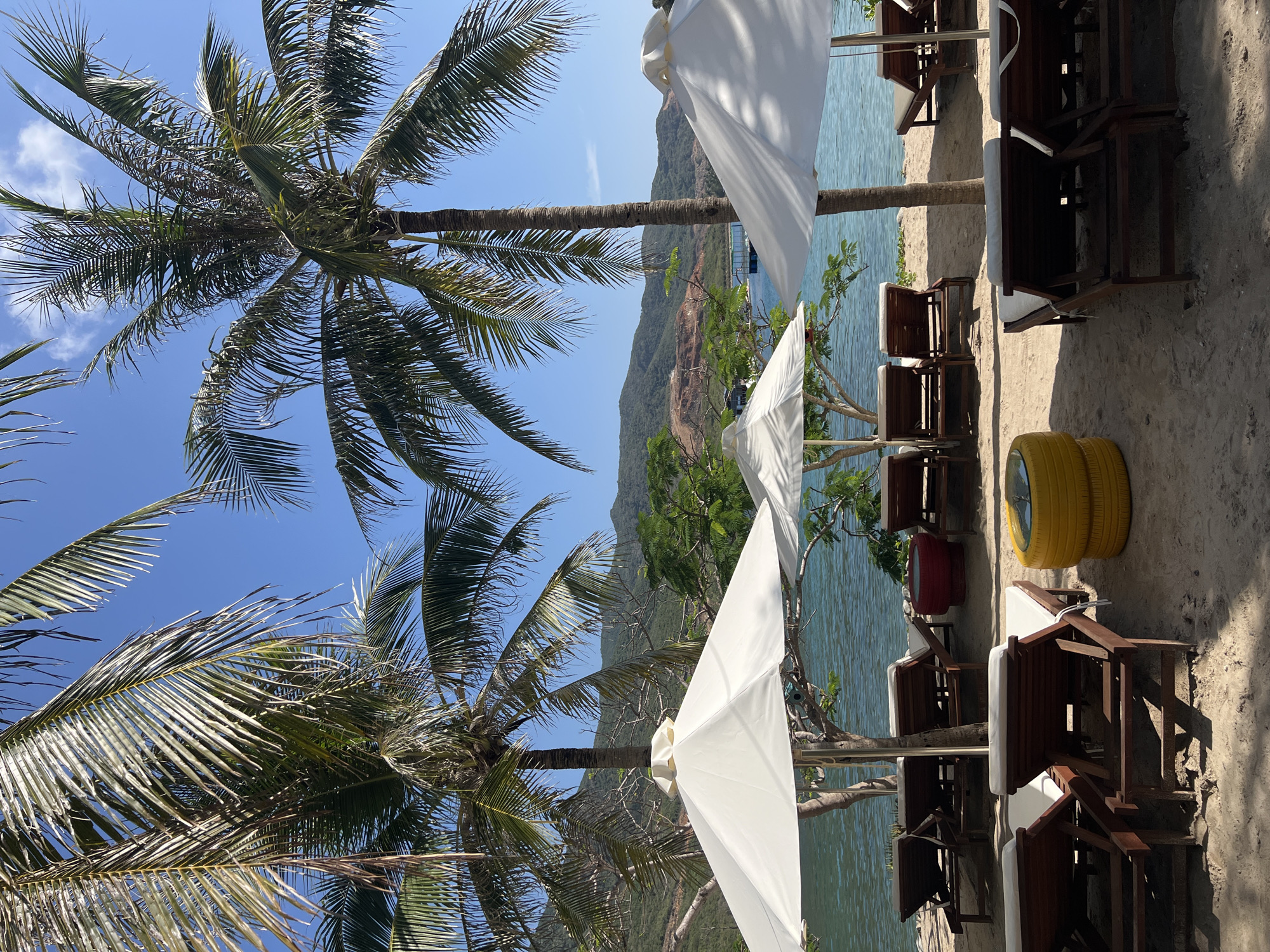 This scene shows a beach in Nha Trang Ward, Vietnam. The area is lined with palm trees and white sun umbrellas. Wooden beach chairs with white cushions are arranged on the sand, accompanied by small tables. In the background, a green, hilly mountain range is visible, and the sea is calm and turquoise. A yellow tire is also visible in the corner.