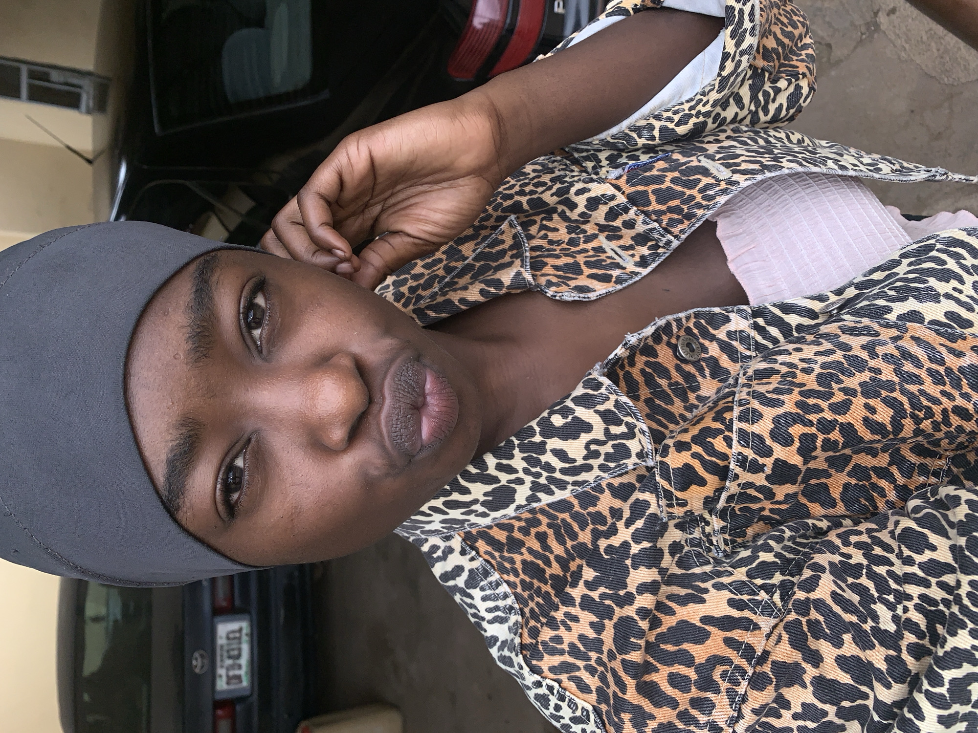A woman is looking directly at the camera, wearing a gray head covering and an open leopard print jacket over a pink top. She has her hand near her ear. The background shows a car, possibly a Toyota, with a license plate partially visible. The location is indicated as Rafin Par, Nigeria. The environment appears to be outdoors, likely near a building.