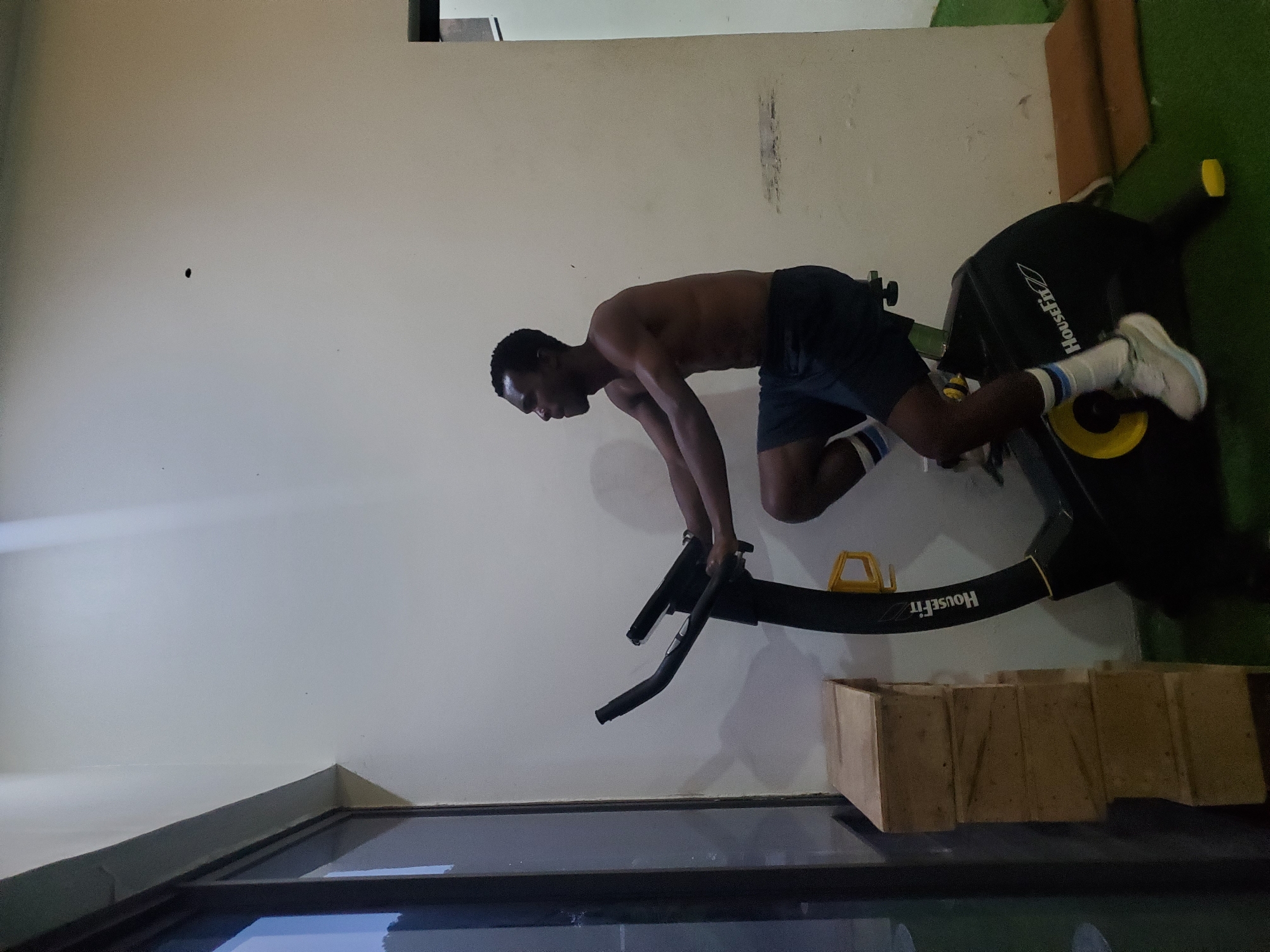 A young Black man is exercising on a stationary bike in a home gym. He is shirtless, wearing dark shorts, white socks, and light-colored sneakers. The bike is black with yellow accents and has "HouseFit" written on it. He is leaning forward, gripping the handlebars, with his body angled as if in motion. The background is a plain, light-colored wall. To the right of the bike, there is a section of green turf, and a few wooden boxes are stacked in the foreground. The lighting suggests it might be indoors during the day or evening. The overall scene is one of focused physical activity.