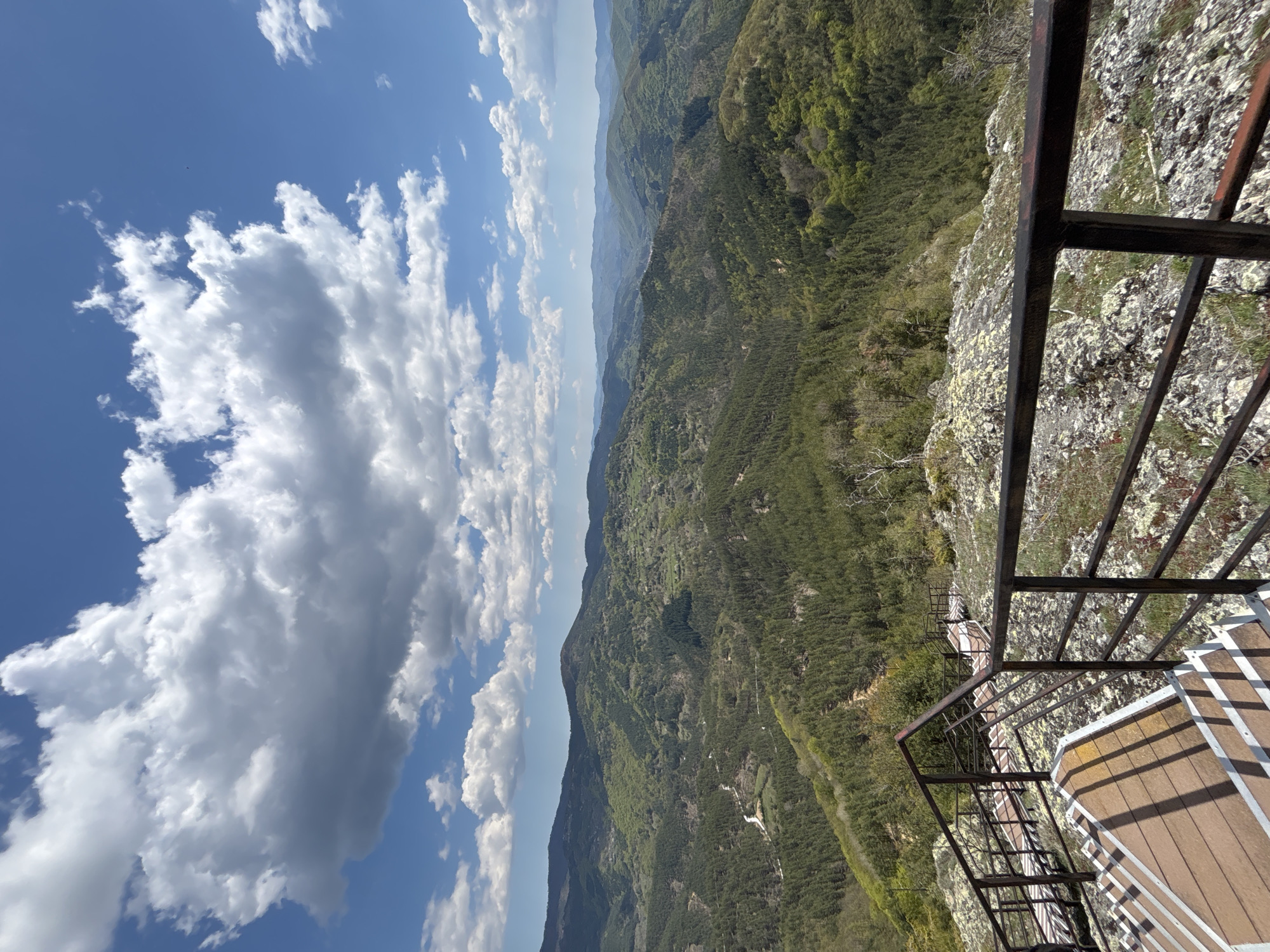 The image shows a view from a high point in Gradat, Bulgaria. The foreground features a metal railing and part of a wooden platform, presumably a viewing point. Below the platform, a steep, rocky cliff descends into a forested valley with layers of distant mountain ranges visible. The sky occupies a significant portion of the image, showcasing a blue expanse with scattered cumulus clouds. There are no people visible.