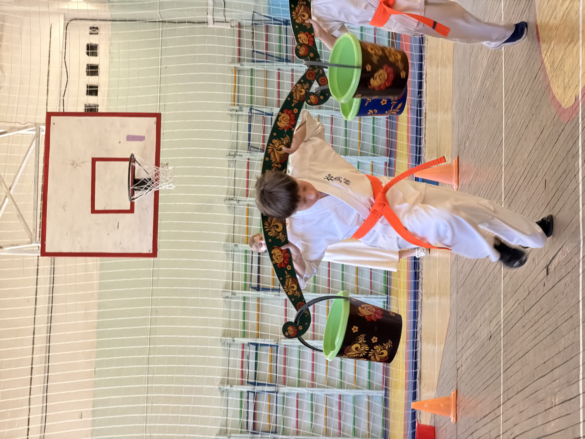 The image shows two people wearing white martial arts uniforms with orange belts, participating in a game in what appears to be a gymnasium. Each person is carrying a yoke with buckets hanging from it. The buckets are dark with floral designs. Orange cones are visible on the floor. A basketball hoop and backboard are mounted on the wall. A safety net is present. The scene is likely in a school or community center in Izhevsk, Russia.