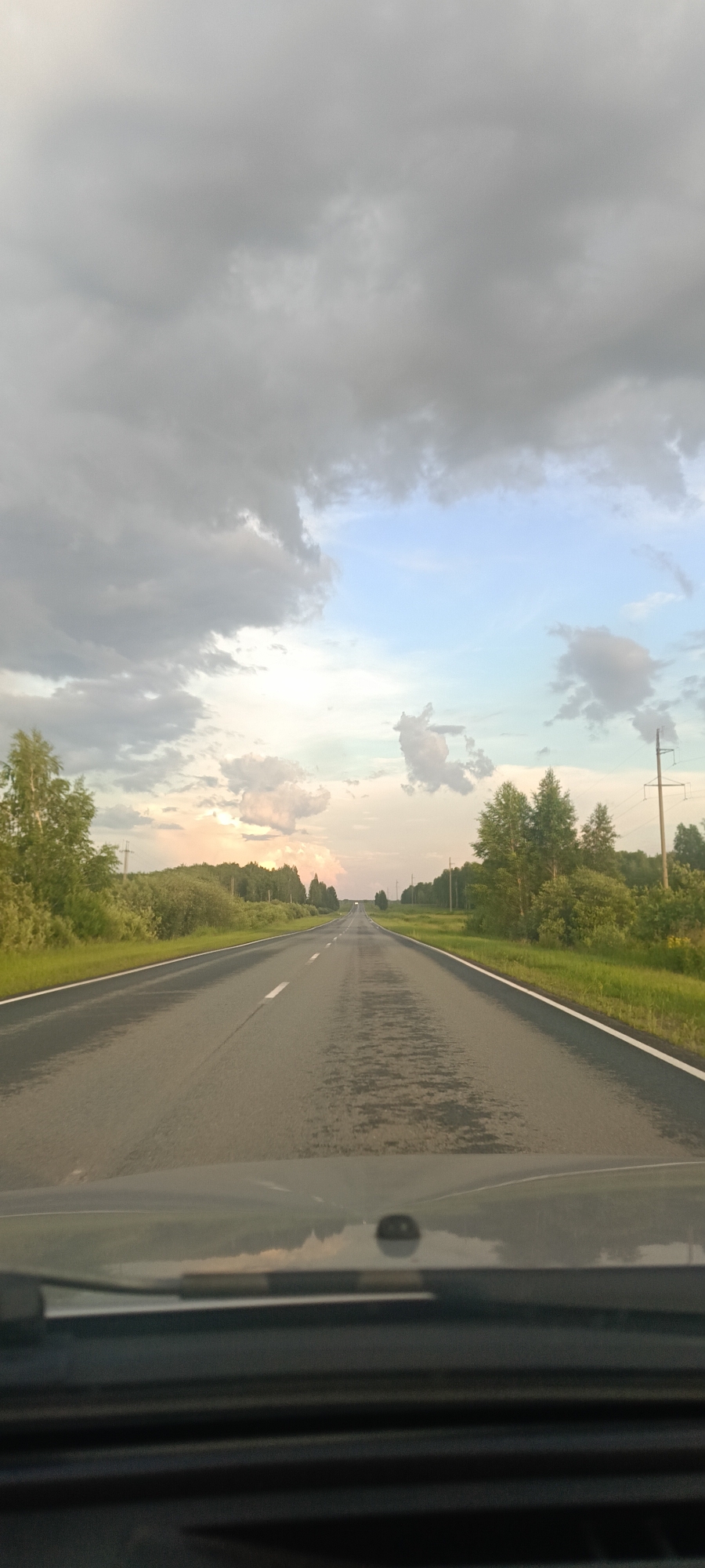 This media file captures a view from the driver's seat of a vehicle traveling on a two-lane asphalt road in a rural setting. The road stretches forward into the distance, flanked by lush green fields and trees on either side. Power lines and utility poles are visible along the roadside, suggesting a semi-developed rural area. The sky above is filled with dramatic, heavy clouds, with patches of lighter sky and hints of sunset colors peeking through on the horizon. The road surface appears wet, indicating recent rain or damp conditions. The overall impression is of a journey through the countryside under a moody, overcast sky, possibly during the late afternoon or early evening. There is no visible text in the image. The scene conveys a sense of travel and perhaps a slight melancholy due to the weather. The location is identified as Malyshevo, Russia.