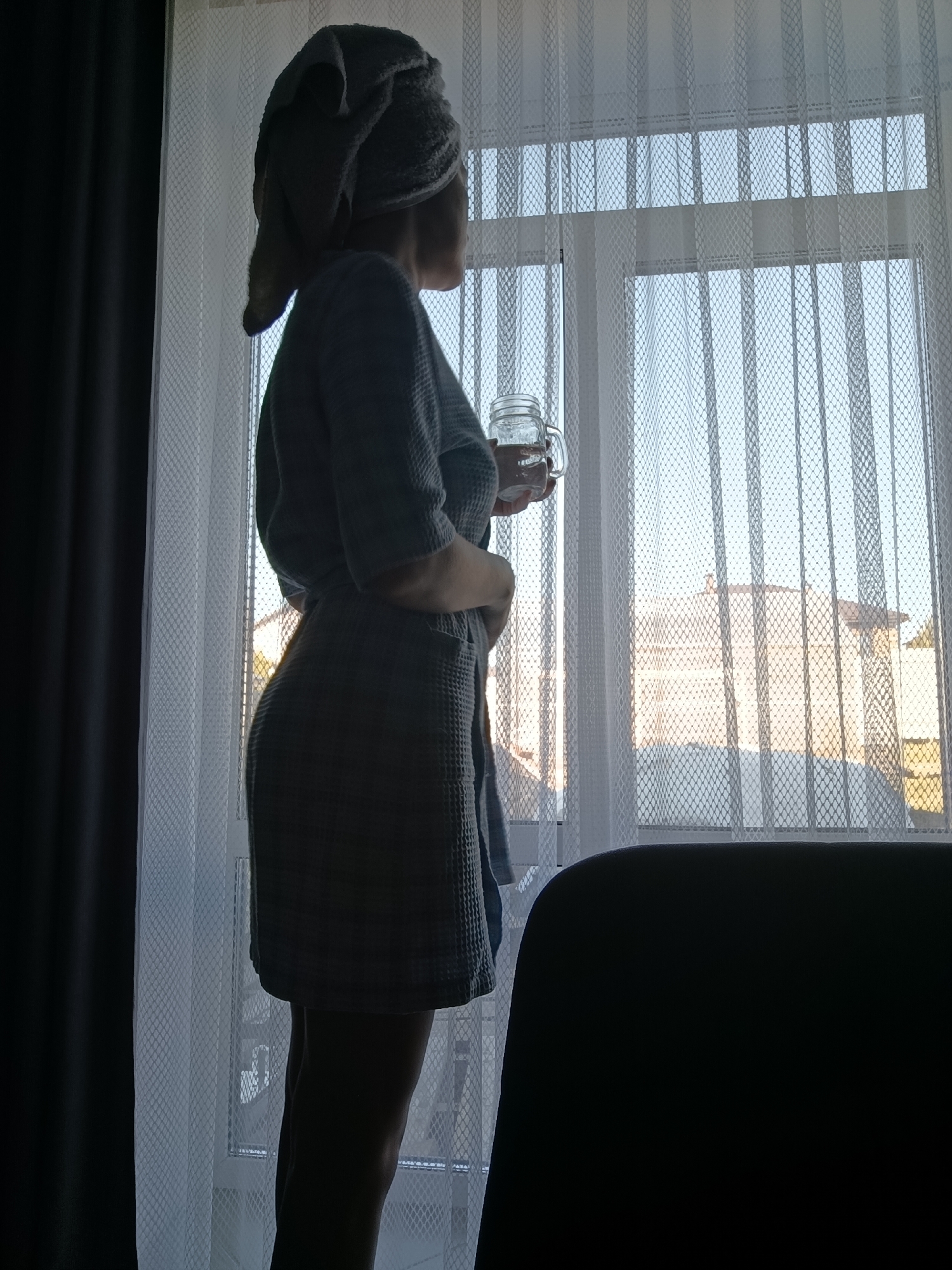 A woman wearing a robe and a towel on her head stands by a window, holding a glass of water. She is facing away from the viewer and looking out the window. The window has white, patterned curtains. A dark curtain is visible on the left side of the frame. Outside the window, buildings can be seen. Based on the information provided, this scene is in the city of Chaikovsky, Russia. A dark object, possibly a chair, is in the foreground on the right.