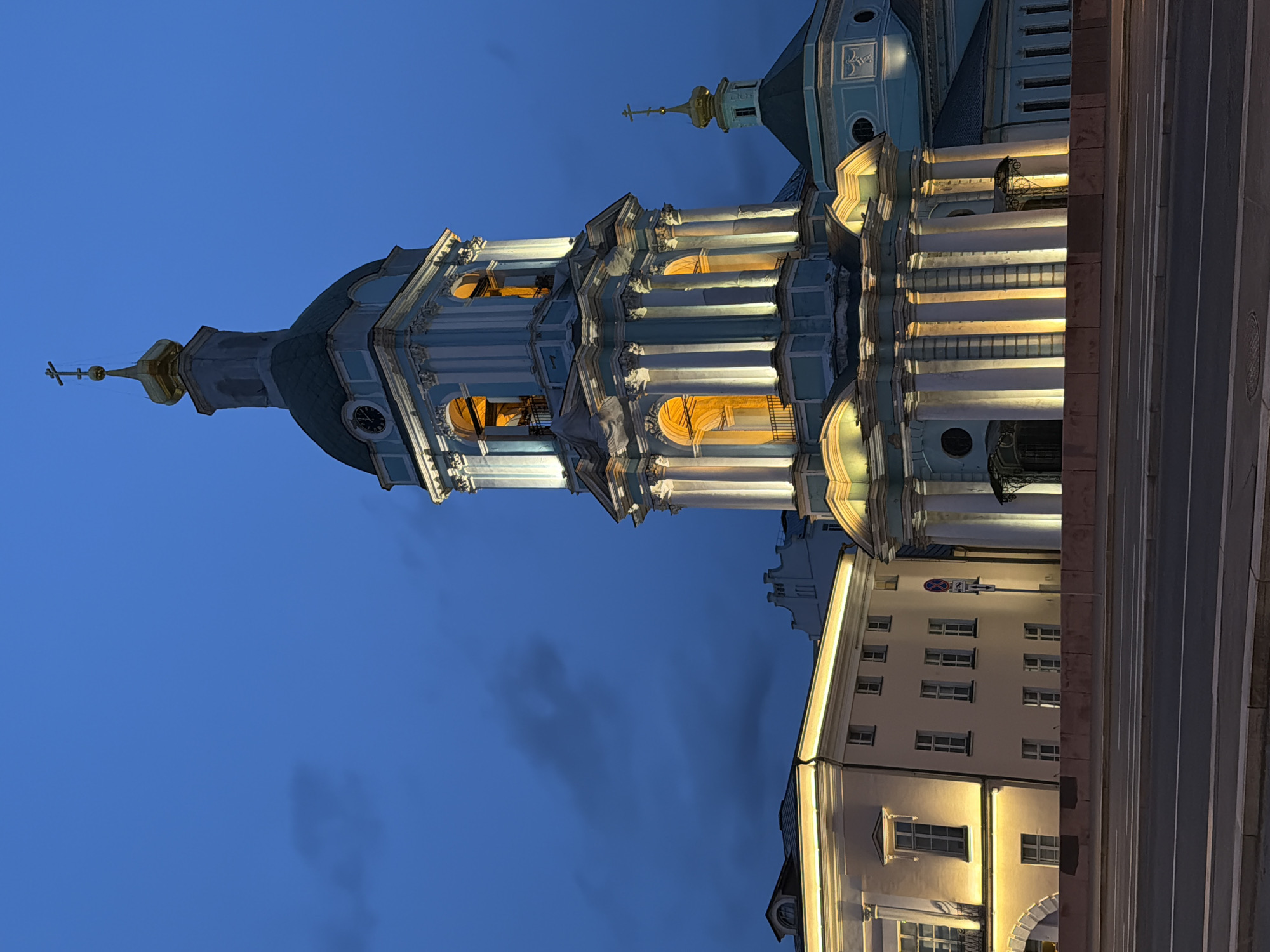 The image shows the bell tower and main dome of St. Nicholas Cathedral in Podolsk, Russia, at dusk or night. The architectural style is Baroque, with blue and white facades and gold accents on the domes and cross. The tower is illuminated with warm light from within, contrasting with the cool blue of the evening sky. Another building is visible beneath the Cathedral. Some road markings are visible in the foreground.