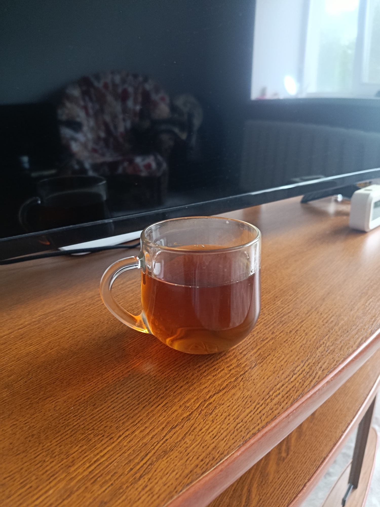 A clear glass mug filled with amber-colored liquid, likely tea, sits on a polished wooden surface. The mug has a curved handle. In the background, a large, dark rectangular object, possibly a television screen, reflects the scene. The screen shows a blurred image of a patterned armchair. Further in the background, a window with natural light is visible. The wooden surface has a prominent grain pattern. The scene appears to be indoors, likely in a living area or office. There is no indication of the city of Syzran, Russia.