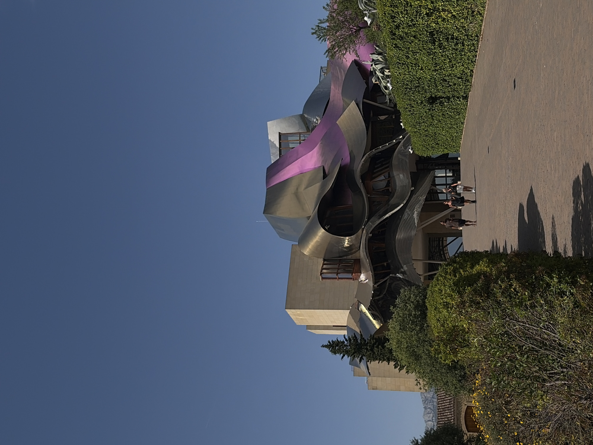 An architectural building is situated on a clear sunny day in Elciego, Spain. The structure features a contemporary design with flowing, metallic surfaces in shades of silver, purple, and black. A traditional building is seen in the background behind the modern building. A paved walkway runs alongside the building, with three people walking in the distance. Landscaping with dense greenery is present along the pathway. The sky is a clear blue.