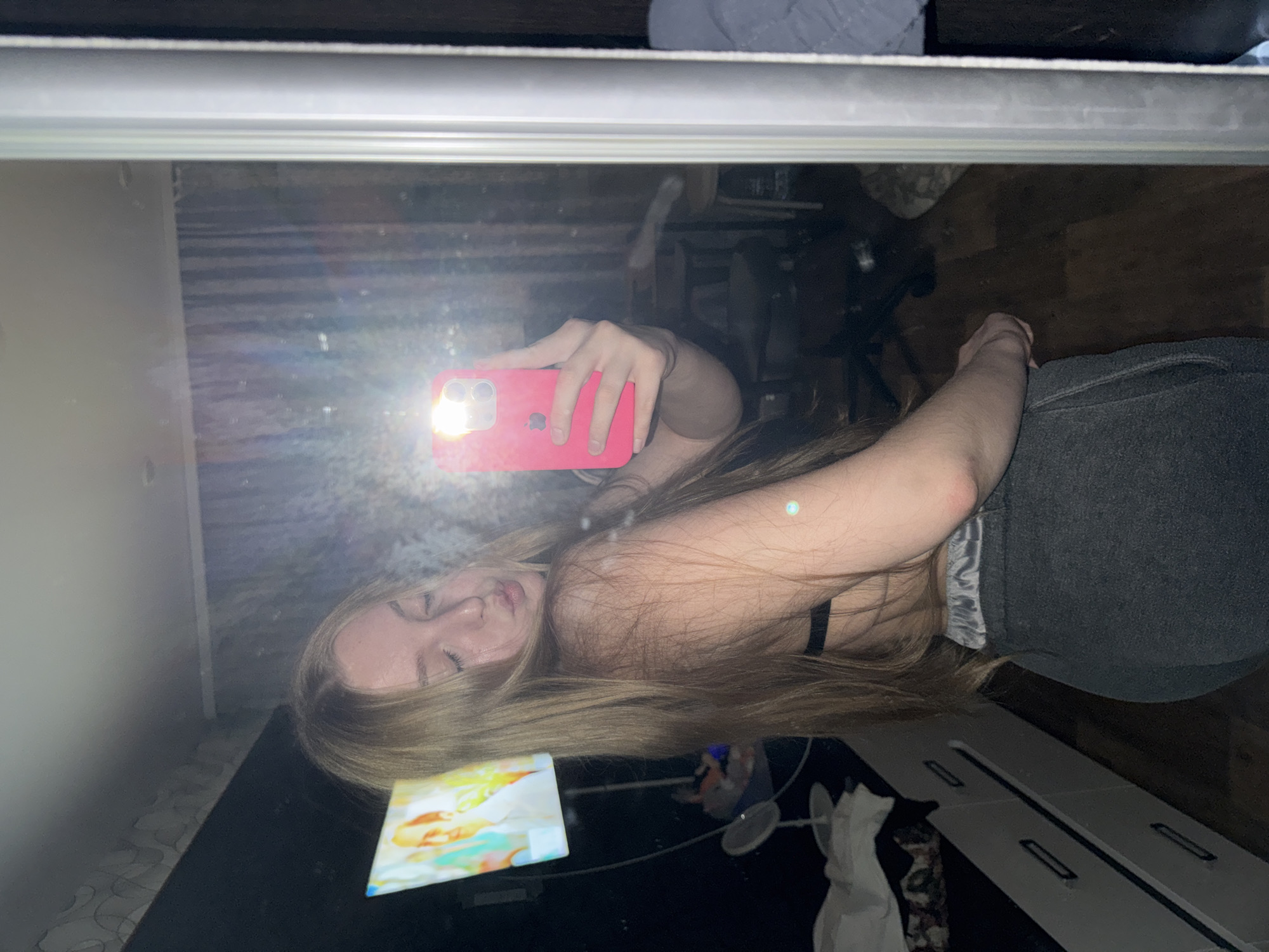 A young Caucasian woman with long blonde hair is taking a mirror selfie in a dimly lit room. She is wearing a black bra and grey sweatpants. She is leaning forward, with her eyes closed and lips pursed as if blowing a kiss. The room appears to be a bedroom, with a dresser on the left and a mirror that takes up most of the right side of the frame. A television screen is visible in the background on the left, displaying an image that appears to be a painting or a still from a movie. There is a pink iPhone in the woman's hand, pointed towards the mirror. The overall impression is a casual, private moment captured at home.