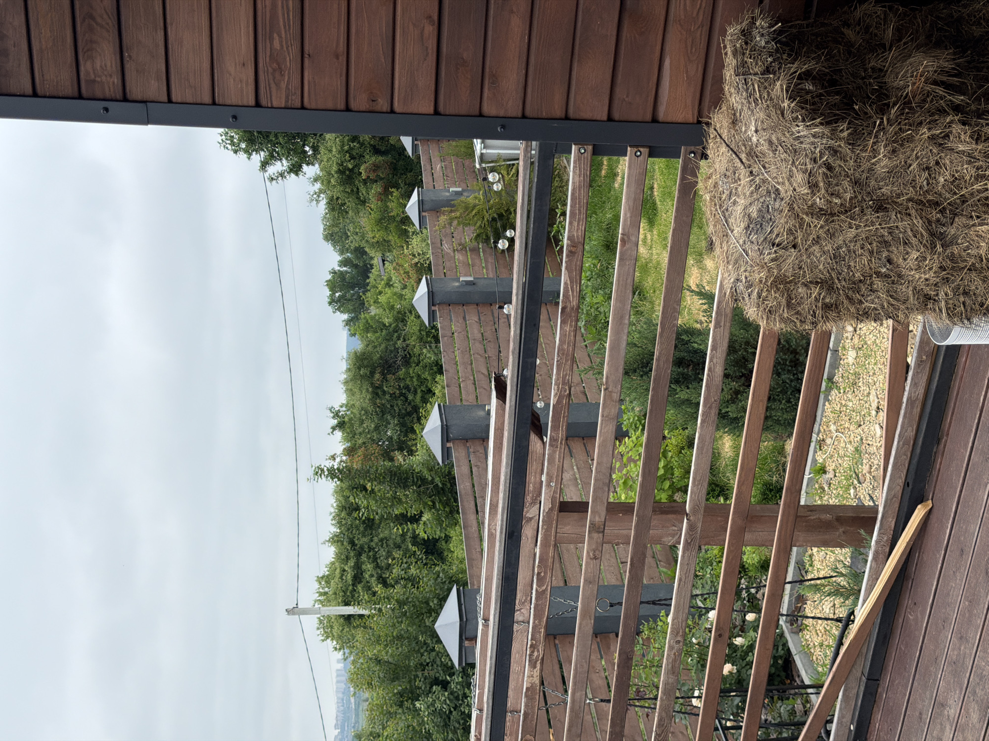 The image shows a view from a wooden deck looking outwards towards a backyard area. In the foreground on the right, a large bale of hay is positioned next to a wooden wall of a building. The deck has wooden planks and a wooden railing with horizontal slats. Beyond the railing, there is a grassy area with some small evergreen trees and white roses. A wooden fence with dark grey posts topped with pyramid-shaped caps runs along the perimeter of the backyard. In the distance, a line of trees and a hazy cityscape are visible under an overcast sky. The overall scene suggests a residential area in or near the city of Semiluki, Russia.