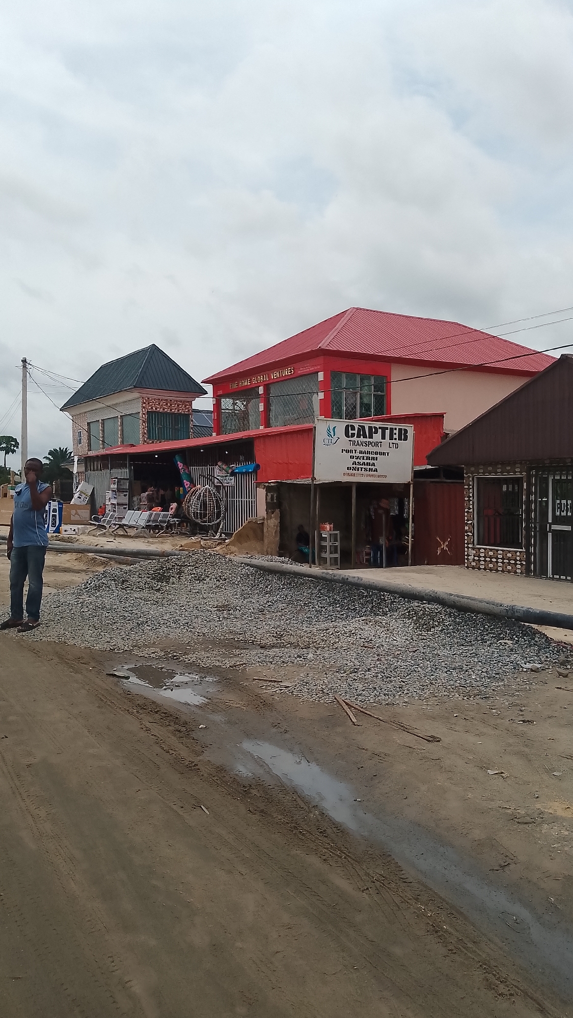 The image depicts a street scene in Unknown, Unknown. In the foreground, there is a dirt road with puddles of water and tire tracks. A pile of gray gravel or small stones is positioned near the edge of the road, close to the buildings lining the street.
On the left side of the frame, a man is standing near the road, wearing a blue shirt and jeans. He appears to be talking on a cell phone.
The buildings in the background include a two-story structure with a dark green roof and decorative brickwork, a red building with a sign that reads "LIVEHOME GLOBAL VENTURES," and a smaller structure with a brown roof. Adjacent to the red building is a sign that reads "CAPTEB TRANSPORT LTD" along with a list of cities: "PORT-HARCOURT, OWERRI, ASABA, ONITSHA." There are also people visible in the open areas of the buildings.
The sky is overcast, with gray clouds covering the majority of the view. Utility poles and wires are also visible above the buildings.