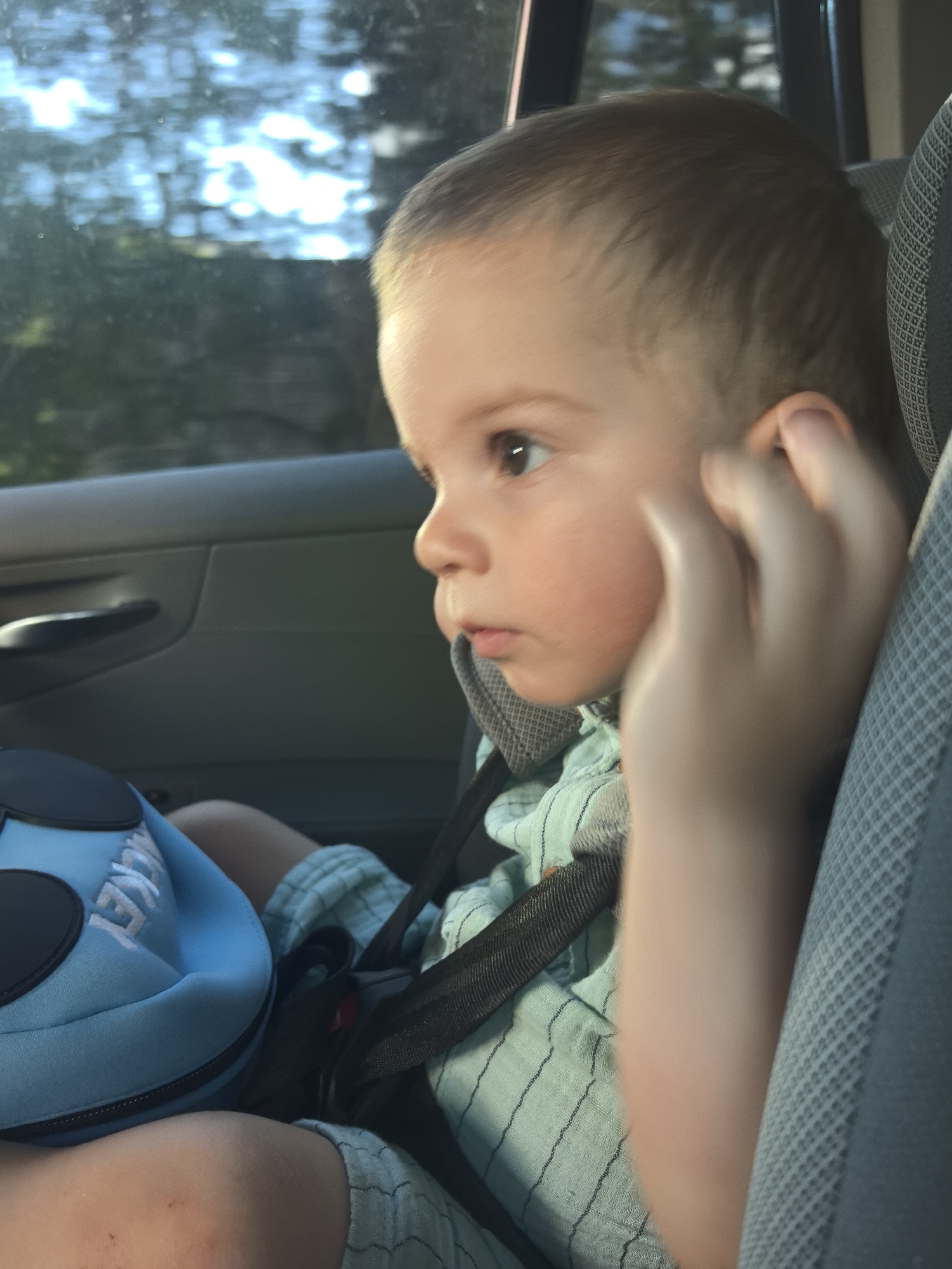 A close-up, eye-level shot captures a young child sitting in a car seat, looking out a window. The child, with light brown hair and fair skin, is wearing a light blue and white striped shirt and is secured by a black car seat harness. Their right hand is raised to their cheek. To the child's left, a blue object with black circles and white lettering that reads "JECKET" is visible. The background shows a blurred view of trees and sky through the car window, illuminated by sunlight. The interior of the car, including the door panel and car seat fabric, is also visible.