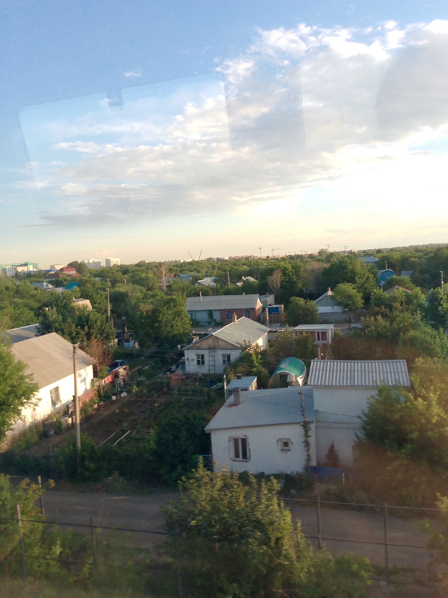 This media file shows an aerial view of a residential area, likely a suburban or semi-rural setting, under a partly cloudy sky. The scene is dominated by numerous houses of varying sizes and architectural styles, interspersed with dense green trees and vegetation. Some houses appear to be single-story dwellings with pitched roofs, while others are more modern, box-like structures. There are also hints of infrastructure, such as power lines and possibly construction cranes in the distance, suggesting ongoing development or a mixed urban-rural landscape.
The time of day appears to be late afternoon or early evening, as indicated by the warm, soft lighting and long shadows cast by the buildings and trees. The sky is a pale blue with scattered white clouds, suggesting fair weather. The presence of lush greenery implies a warm season, possibly summer or late spring.
The image is captured from an elevated position, likely from a window or a drone, looking down and across the landscape. There are no visible people in the image. The overall impression is one of a tranquil, residential community situated amidst nature. The quality of the image is somewhat grainy, and there are some reflections or imperfections visible on what appears to be a glass surface through which the photo was taken, particularly in the upper portion of the image showing the sky. No text is visible in the image.