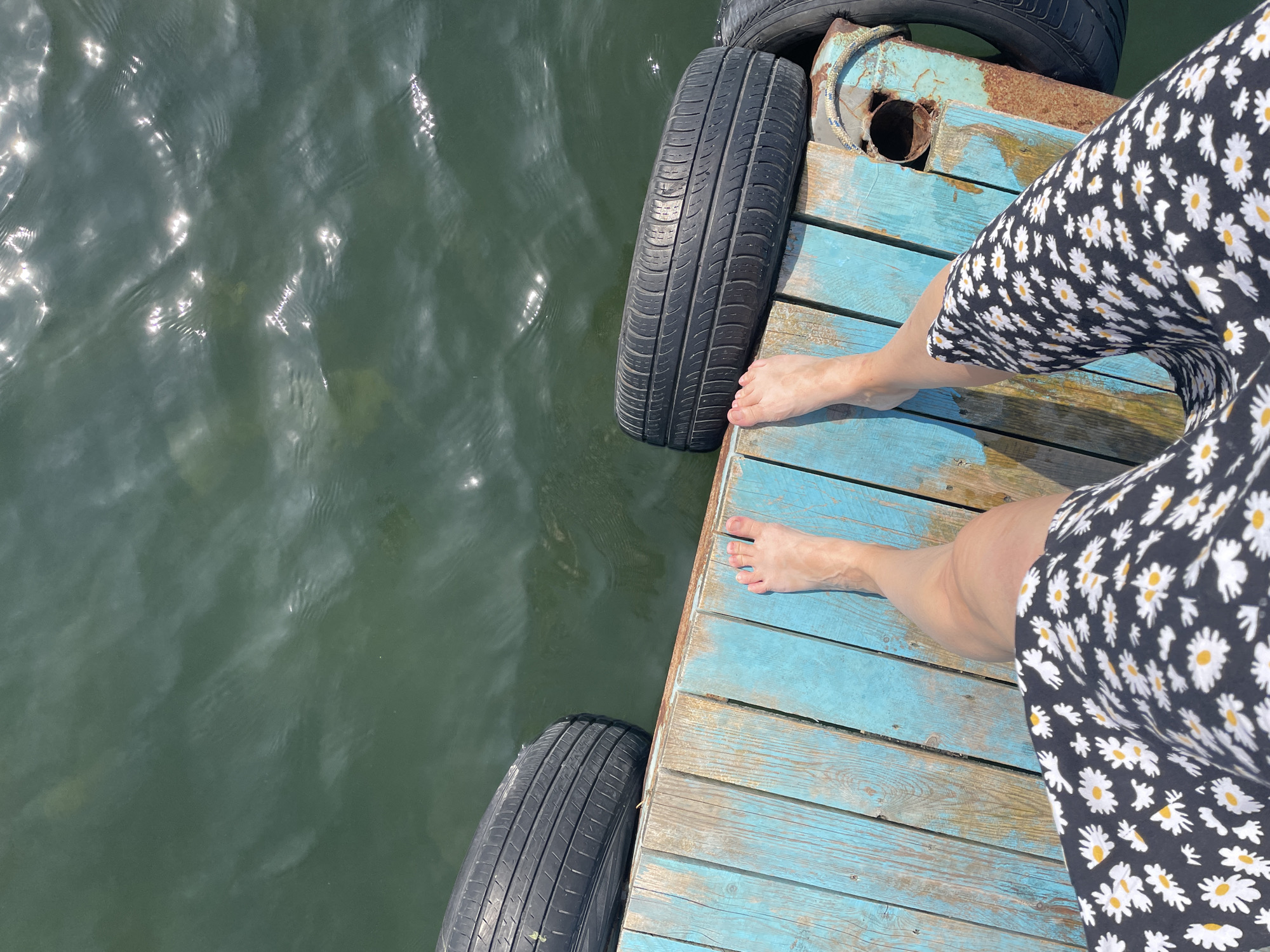 A person's bare feet are visible on a weathered, blue wooden dock. The person is wearing a black dress with a white daisy pattern. Two tires are positioned on the edge of the dock, one to the left and another to the right, partially submerged in dark, rippling water. Sunlight reflects off the water's surface. The scene is likely outdoors by a body of water.