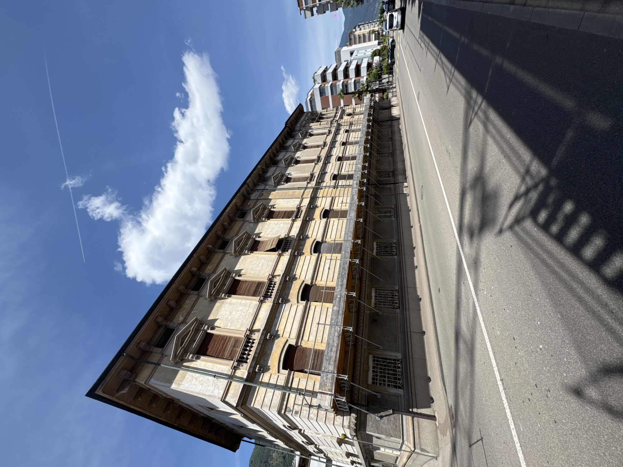 The image shows a street in Locarno, Switzerland, with a long, historic building on the left and modern buildings across the street on the right. The historic building is a light beige color with ornate architectural details around the windows and a dark-colored roof. Scaffolding is erected along the lower part of the building. The street in front of the building is paved and has a white dividing line. A shadow of intricate ironwork is cast across the road. Several cars are visible on the street, including a white van and a dark-colored sedan. In the background, there are mountains and a bright blue sky with scattered white clouds and a contrail from an airplane.