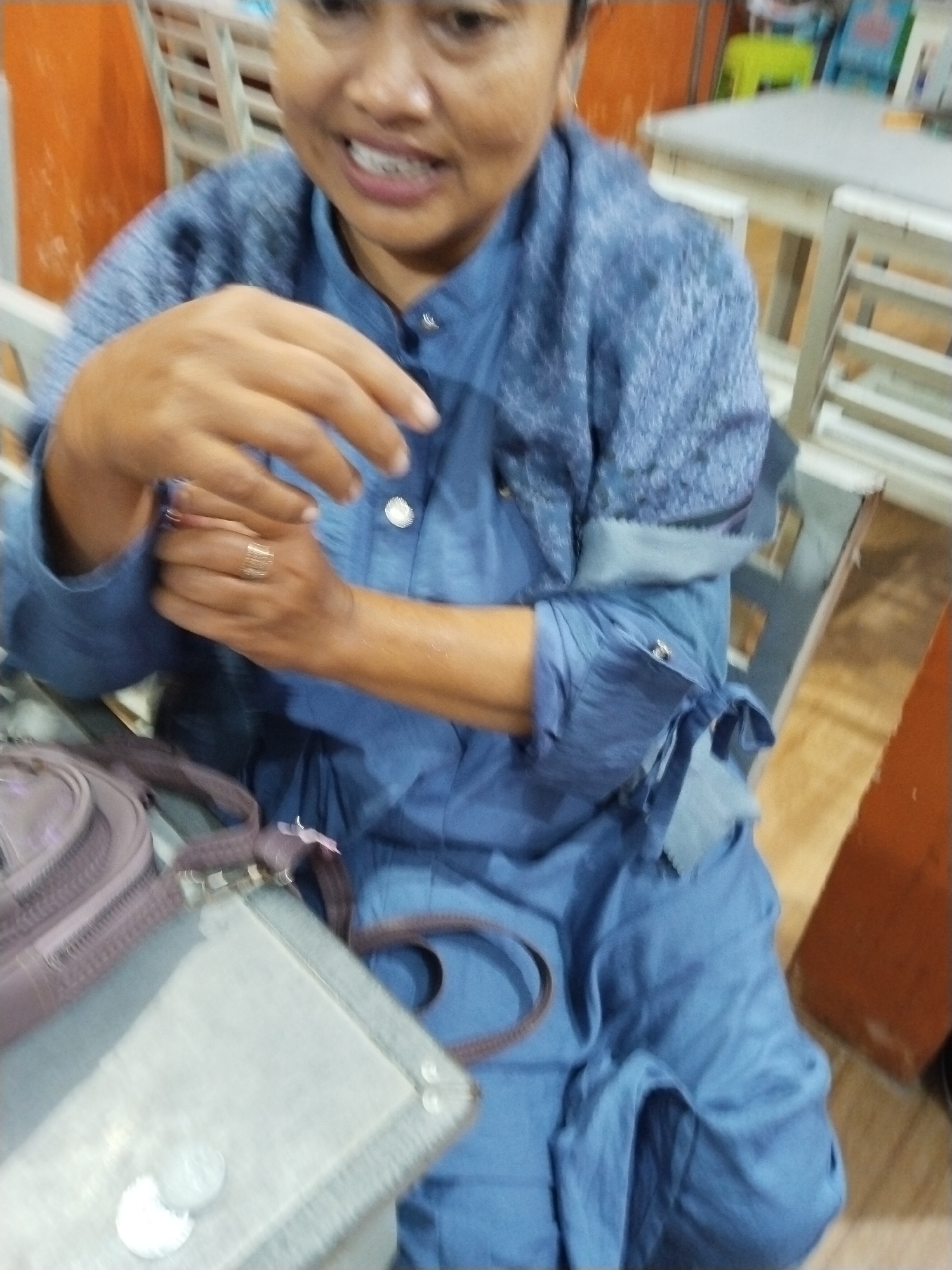 A medium shot shows a woman in a blue tunic with a textured yoke and cuffs. She is seated at a table, with her hands clasped in front of her. A ring with multiple bands is visible on her left ring finger. A brown handbag with a purple strap is resting on a light-colored surface in the foreground, next to two coins. The background is blurred, showing glimpses of furniture and an orange wall. The overall impression is that of an indoor scene, possibly a cafe or restaurant, in Prigi, Indonesia.