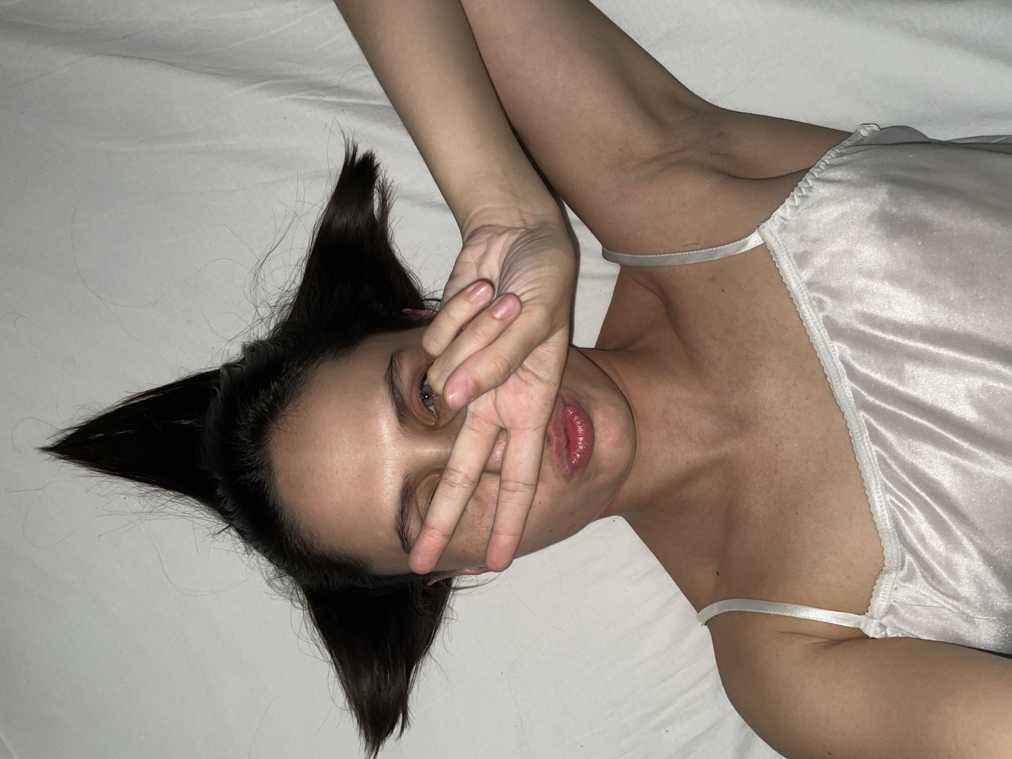 A woman lies on her back on a white surface, possibly a bed, wearing a white top with thin straps. Her dark hair is styled into sharp, angled points, resembling cat ears. She has one hand raised, covering part of her face. Her eyes are visible, and her lips are slightly parted. The lighting appears to be dim. The location in Izhevsk, Russia, is not discernible from the image.