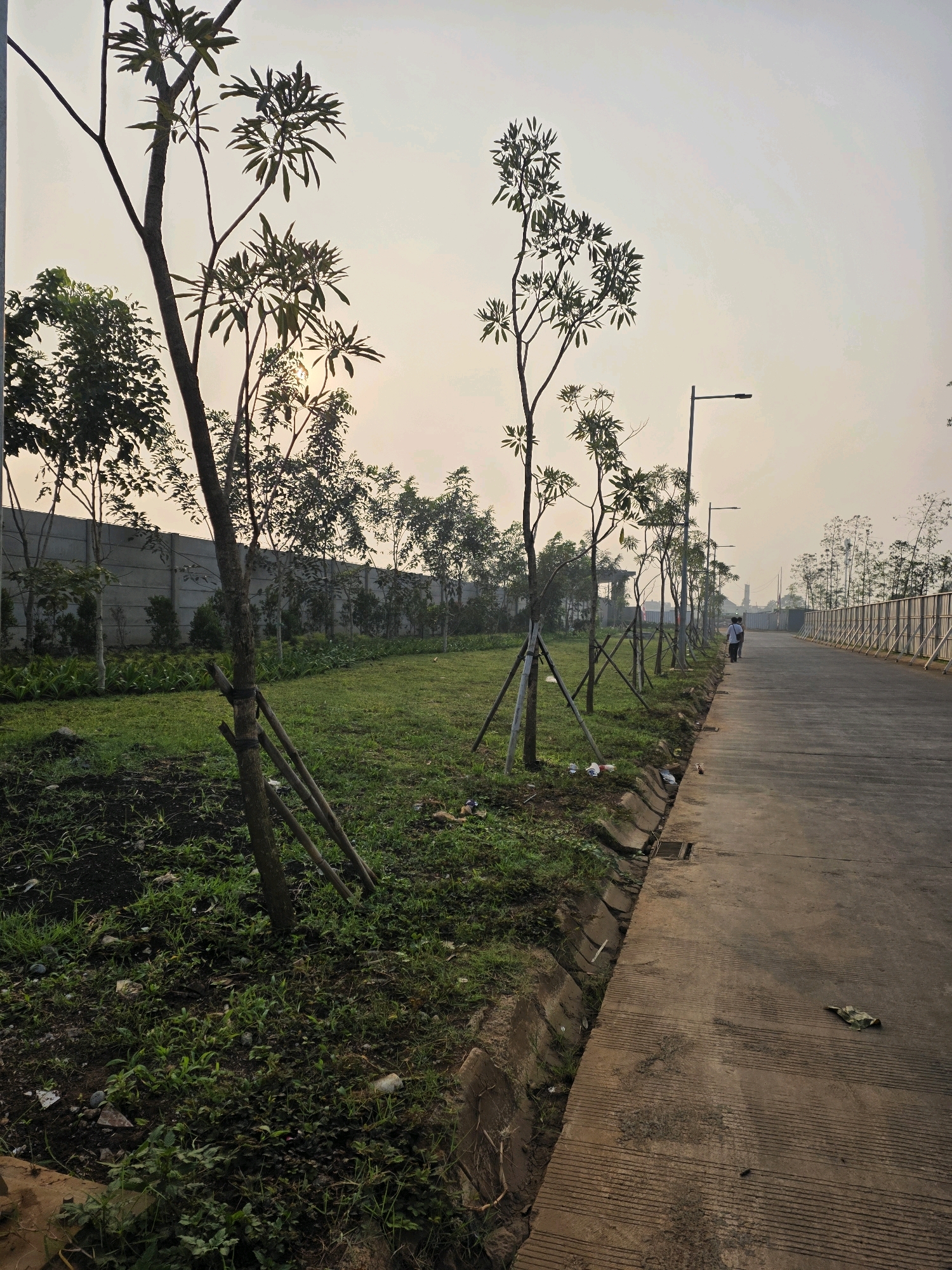 In Bojongsoang, Indonesia, a paved road stretches into the distance, bordered on the left by a grassy area with young trees staked to the ground. A concrete wall runs along the far side of the grassy area, and beyond that, more trees are visible. Several streetlights line the right side of the road. Two individuals are walking away from the camera on the road. The sky is overcast, with the sun faintly visible through the haze.