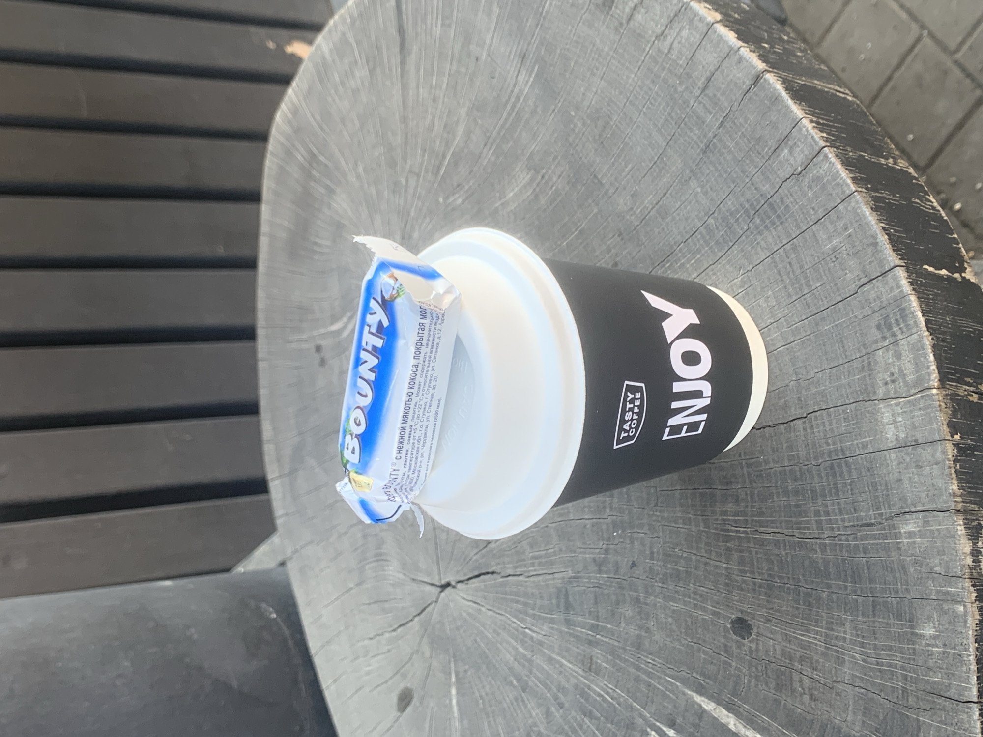 The image shows a partially eaten Bounty chocolate bar resting on the lid of a black and white coffee cup labeled "Tasty Coffee" and "ENJOY". The cup sits on a weathered, round wooden surface resembling a tree stump. The surface is likely part of a table or seating area. In the background, there's a glimpse of a dark gray bench with horizontal slats. The scene is captured outdoors, possibly in a park or urban area. Based on the text in the chocolate bar packaging, the image likely originates from or depicts a location within Russia. The text mentions production details related to Stupino, Moscow Region, and Cherdakly, Ulyanovsk region. However, there is no indication of the exact location as Izhevsk.
