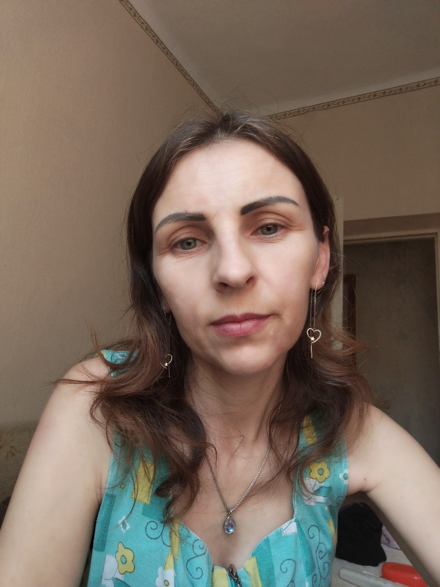 A head-and-shoulders, front-facing medium shot captures a woman indoors, seated and looking directly at the camera. She has brown hair, parted and falling around her shoulders, with bangs brushed to the side. Her eyes are green, and she has defined eyebrows. She is wearing a teal tank top adorned with a pattern of yellow and white flowers. A silver necklace with a teardrop-shaped blue pendant rests against her décolletage. Dangling from her right ear is a long silver earring with a heart-shaped charm and a small star. The background consists of a plain, light brown wall, with a decorative border running along the top. To the right, a partially visible doorway leads to a dimly lit room. The lighting suggests an indoor setting, possibly a home. The image was taken in Vinnytsia, Ukraine.