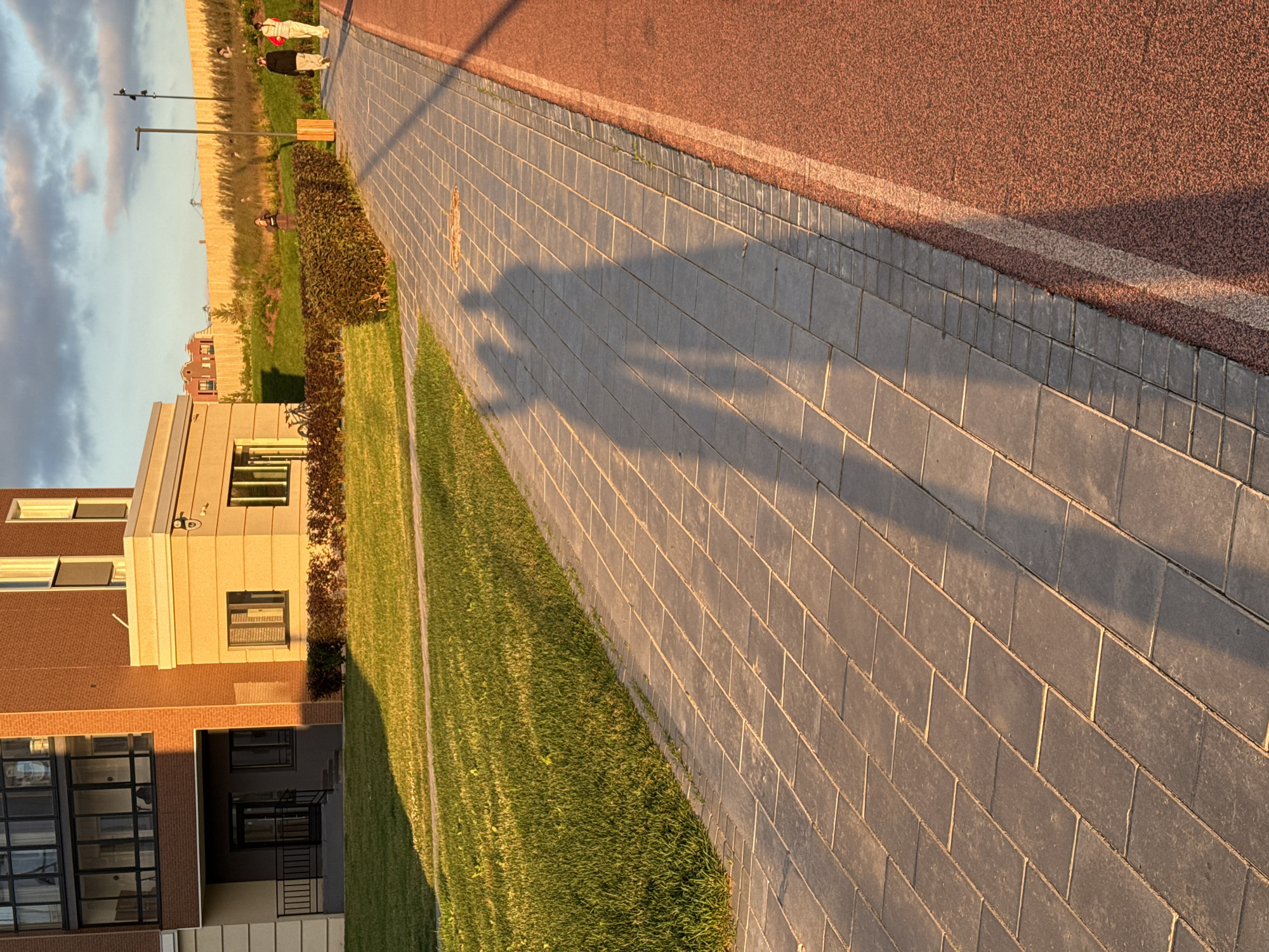 The image depicts an outdoor scene, likely in a modern urban or suburban environment, possibly Pervomaysky, Russia. The perspective is from a low angle, looking across a paved walkway made of grey rectangular tiles. To the left of the walkway is a neatly trimmed grassy area, and further left, a contemporary building with a brick facade and large windows. Beyond the grass and walkway, a reddish-brown textured surface, possibly a running track or sports court, is visible. The sky above is partly cloudy, with the warm light suggesting it is either early morning or late afternoon.
A prominent feature of the image is the long shadow cast across the paved walkway. This shadow appears to be from two people, likely walking or standing together, though they are not directly visible in the frame. Their forms are elongated and distorted by the low-angle sunlight.
In the background, partially obscured by vegetation, is a light-colored building with a distinctive architectural style, featuring vertical elements and windows. Further in the distance, a fence and trees can be seen, bordering a flatter, lighter colored area that might be a field or another part of the urban landscape.
Notable details include the contrast between the hard textures of the pavement and the reddish surface and the soft textures of the grass and foliage. The golden hour lighting creates long shadows and highlights the different surfaces, giving the scene a serene and tranquil atmosphere. No visible text is present in the image. The activity taking place is implied to be leisurely, with people enjoying the outdoor space.