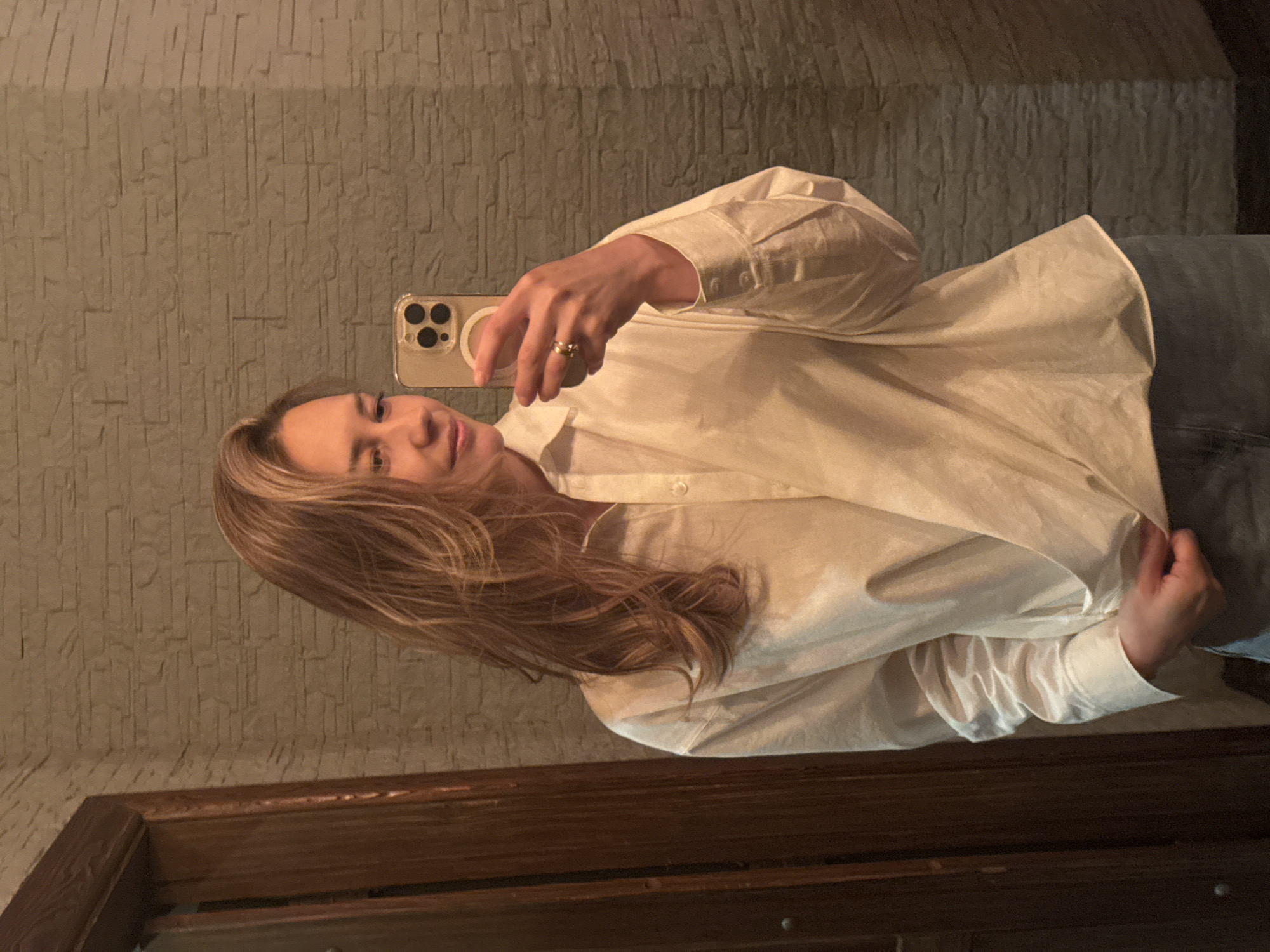 A woman is taking a selfie in front of a mirror. She is wearing a white oversized button-down shirt and jeans. She is holding a phone with a clear case and two visible camera lenses. Her hair is long and blonde. The background is a textured wall and a dark wooden frame, possibly a door or mirror frame. The location is likely indoors but there is no further information suggesting it is in the city of Chaikovsky, Russia.
