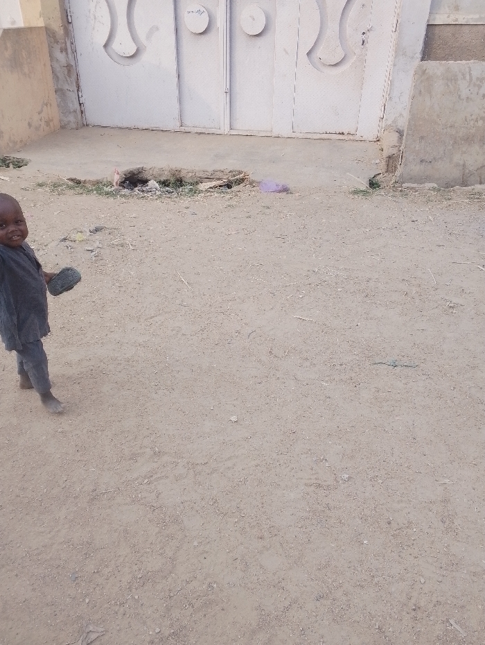 A young child stands barefoot in a sandy area in front of a white metal gate. The child is wearing a dark outfit and holds something in one hand. A small, circular pile of debris and litter sits on the ground near the gate, with a glimpse of greenery. The background shows portions of a beige building. The area appears arid.