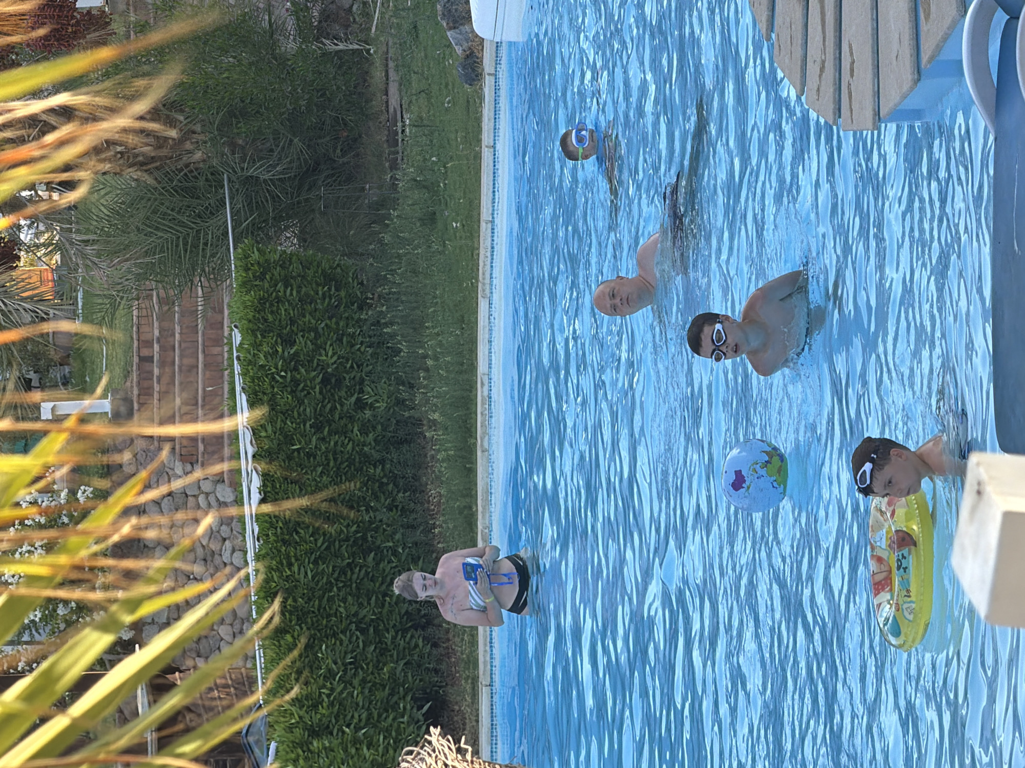 In a refreshing swimming pool in Sharm El Sheikh, Egypt, several people are enjoying a sunny day. A young boy, wearing goggles, is visible near a yellow inflatable ring with cartoon characters. A younger child, also with goggles, is in the water, partially submerged. Further in the pool, another boy, wearing a blue and white striped shirt and black shorts, is standing. A person, possibly a young woman, is seen in the shallow end of the pool, holding a rectangular object with a blue frame, possibly a tablet or waterproof camera. The water is clear and blue, reflecting the bright sunlight. Surrounding the pool are lush green trees and some stone pathways, suggesting a tropical or resort setting. The overall scene conveys a sense of leisure and fun, typical of a vacation day at a pool. The time of day appears to be daytime, likely late morning or afternoon, given the strong sunlight and clear sky.