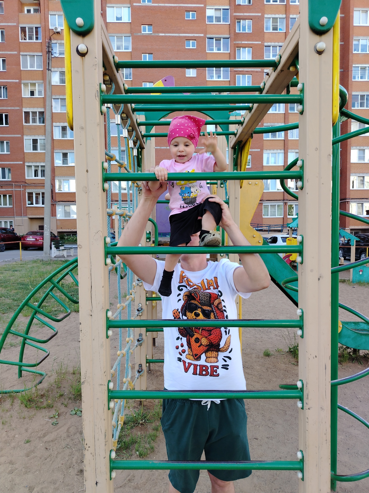 In Chaikovsky, Russia, a father lifts his young daughter atop a playground ladder. The child, wearing a pink hat and t-shirt, holds onto the green bars with one hand raised. The father, in a white t-shirt with a cartoon graphic and dark shorts, supports her from below. The playground is situated in front of a tall brick apartment building with numerous windows. A climbing structure and slide are visible in the background, along with parked cars. The ground is a mix of sand and grass.