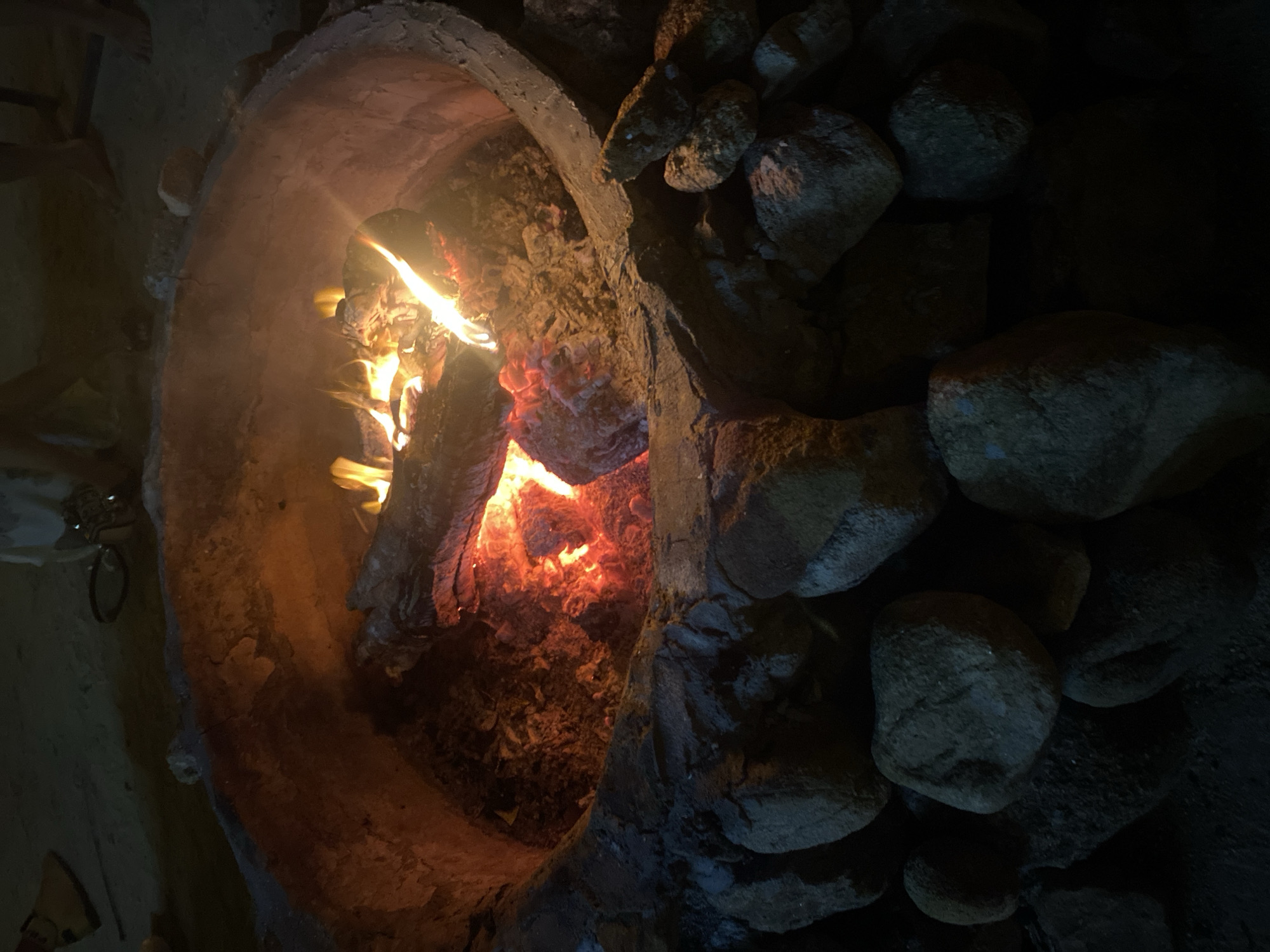 The image depicts a fire burning inside a cylindrical, concrete or stone structure. The structure is constructed using rocks and what seems to be concrete. Flames and burning logs are visible inside the structure, providing illumination. The surrounding area is dark, with more rocks piled to the right of the structure. Based on the metadata, the location of the image is Baan Hin Kong, Thailand. There are no visible people in the image.