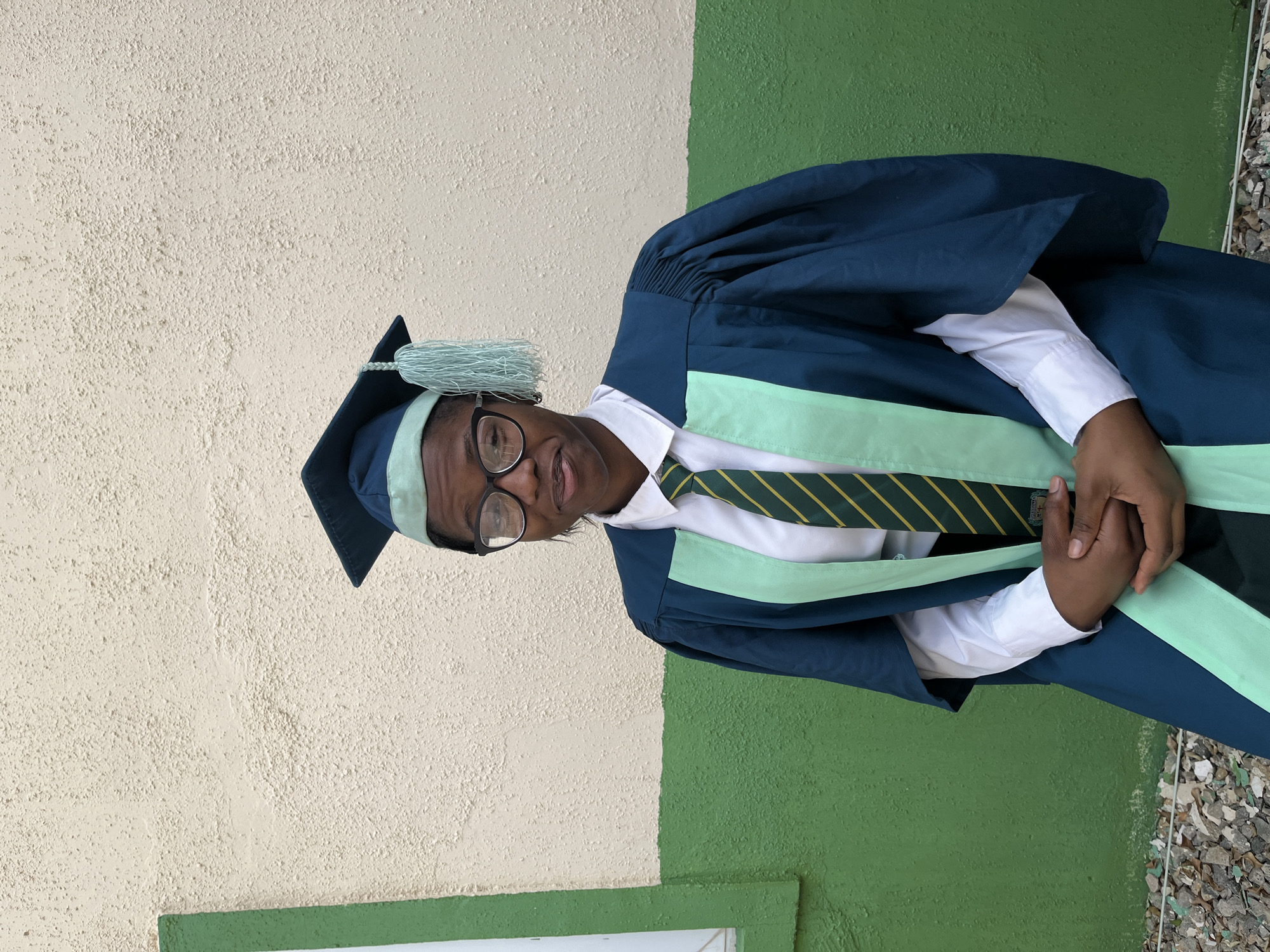 A young person is wearing a graduation gown, cap, and tie. The gown is dark blue with light green accents. The cap is dark blue with a light green tassel. They are wearing glasses. The background is a wall that is painted half cream and half green. The picture appears to be taken outside in a setting consistent with a residential or institutional building. The presence of a graduation gown, cap, and tie suggests the person is likely in a place affiliated with an educational institution, but the exact location within the city of Jos, Nigeria, cannot be determined from the image.