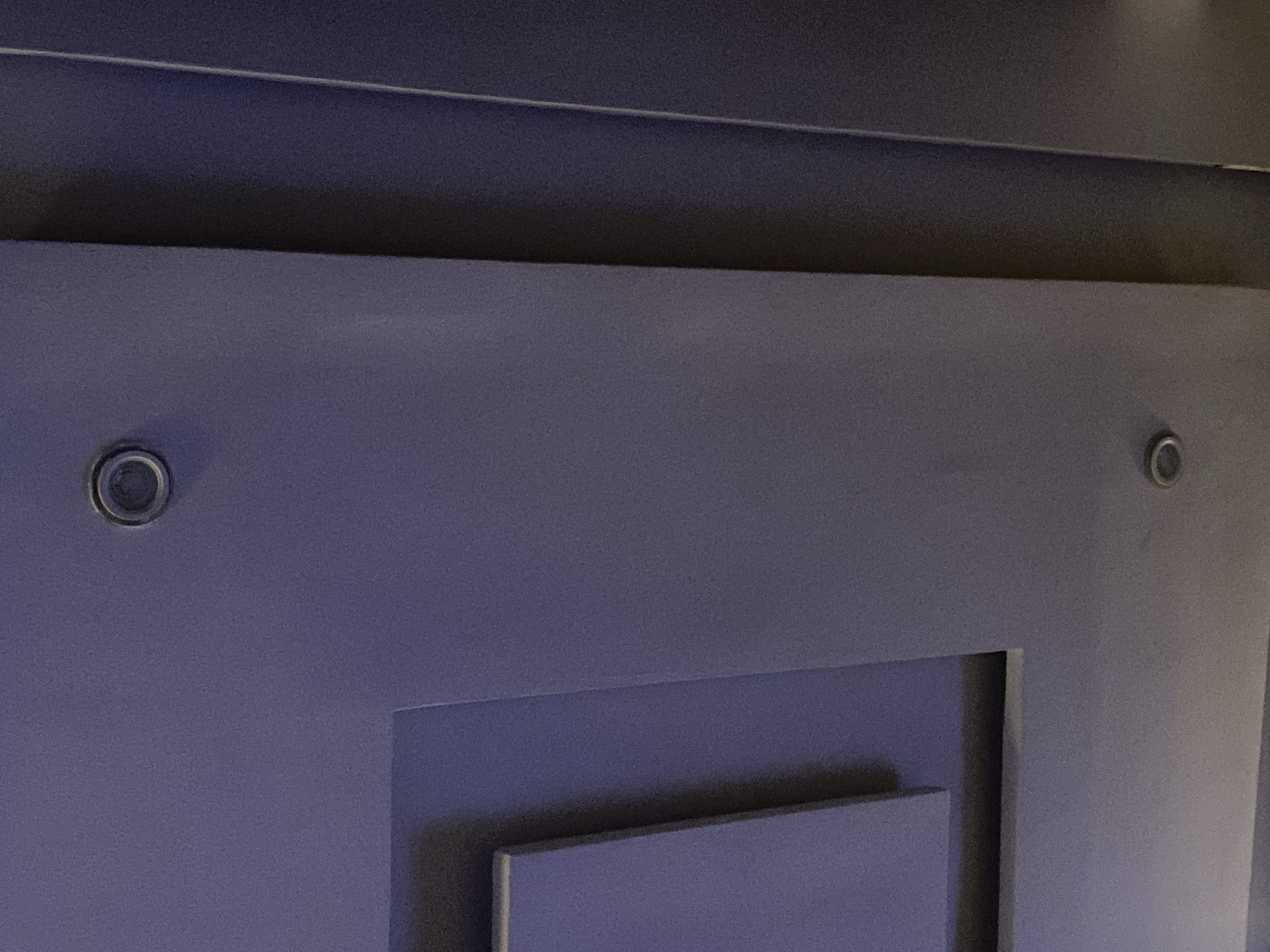 The image shows a close-up view of a modern ceiling with recessed lighting. The ceiling is painted a dark gray or purple hue and features a multi-layered design with geometric shapes. Two circular recessed lights are visible, one near the top right and another near the bottom center of the frame. A vertical dark band runs along the right side of the image, suggesting a transition to another architectural element or a wall. The lighting is subdued, casting soft shadows and highlighting the textured surface of the ceiling. There are no people or any other discernible objects in the image. The location of Giginyu, Nigeria is not determinable from the visual information provided.