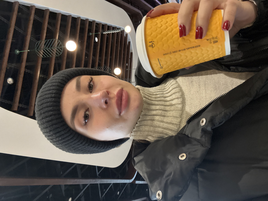 The image displays a light-skinned female in an indoor setting, identified as a McDonald's restaurant, specifically resembling the one at Vokzalna Square in Kyiv, Ukraine, based on the distinctive ceiling architecture. The subject, captured in a selfie perspective from the chest up, wears a black ribbed beanie, a light beige ribbed turtleneck sweater, and a black puffer jacket. Her fingernails are painted dark red. She holds a yellow, textured McDonald's paper coffee cup with a white lid in her right hand; visible markings include "0.2 L" and "© 2020 McDonald's." The background ceiling features a contemporary design with dark wooden beams, white curved panels, and various light fixtures, including spherical pendants and a decorative metal leaf-shaped fixture.