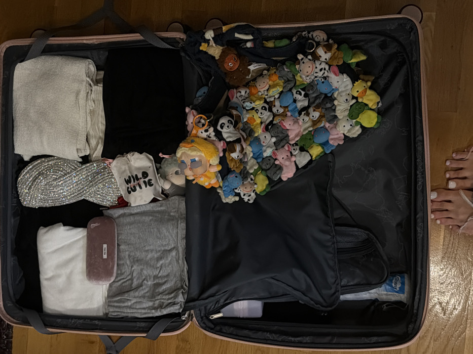 An open, light pink hardshell suitcase is centrally positioned on a light brown wooden floor in Budva, Montenegro. The suitcase is packed with assorted items.
In the upper left compartment, a stack of folded white textile items is visible, alongside a pink velvet eyeglass case branded "Miu Miu," resting on a folded grey t-shirt. A black zippered internal divider covers the lower left section, partially revealing a white plastic bottle underneath.
The upper right compartment contains a folded silver sequined textile item, a folded light beige textured garment, and a small white drawstring bag printed with "WILD CUTIE." Below these items, a dense cluster of numerous small, multi-colored plush animal finger puppets, including pigs, cows, pandas, and frogs, along with a yellow-haired doll-like figure wearing glasses, are contained within a dark mesh bag. Dark-colored garments are also visible within the right compartment.
At the bottom of the frame, a pair of bare human feet with white painted toenails are visible on the wooden floor, positioned as if standing directly over the open suitcase.