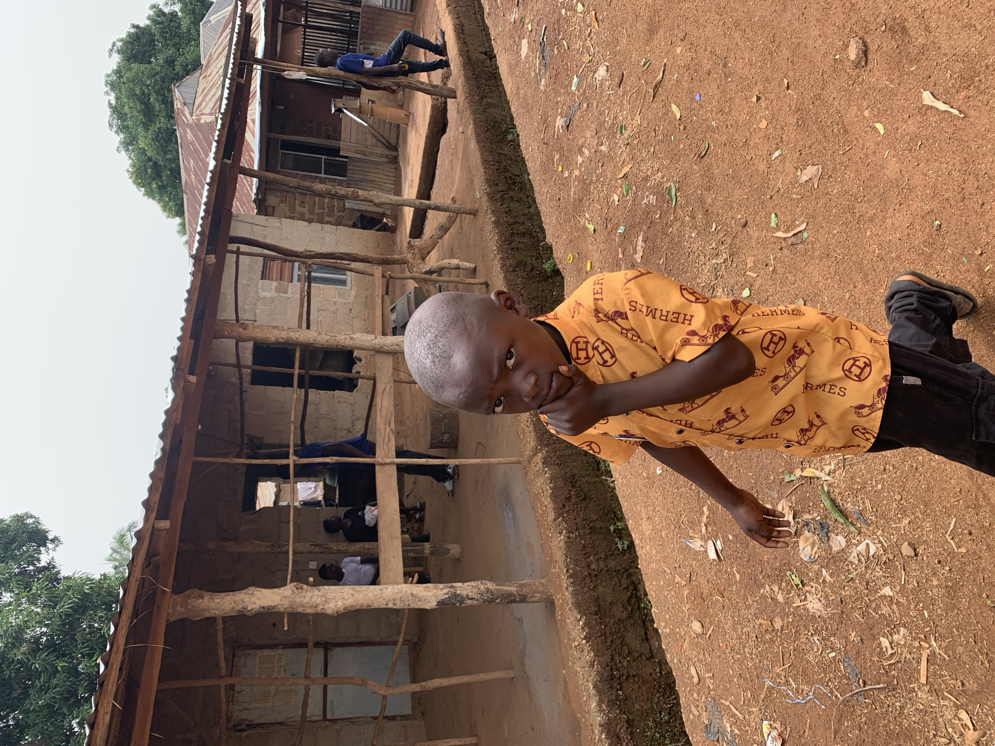 A young boy with a shaved head is the central focus of the photograph. He is wearing an orange shirt patterned with "Hermes" logos. The boy's right hand is raised to his face, resting near his chin. He wears black pants and dark shoes.
Behind the boy is a partially constructed or dilapidated building with a corrugated metal roof. The structure is primarily built of wood and cement blocks. Inside the building, several people are seated. A man is lying on the roof of the building.
The ground is a mixture of reddish-brown soil littered with leaves, debris, and trash. Based on the landscape and architecture, the location appears to be a rural or developing area. Given the reference, the location is purportedly Agban, Nigeria.