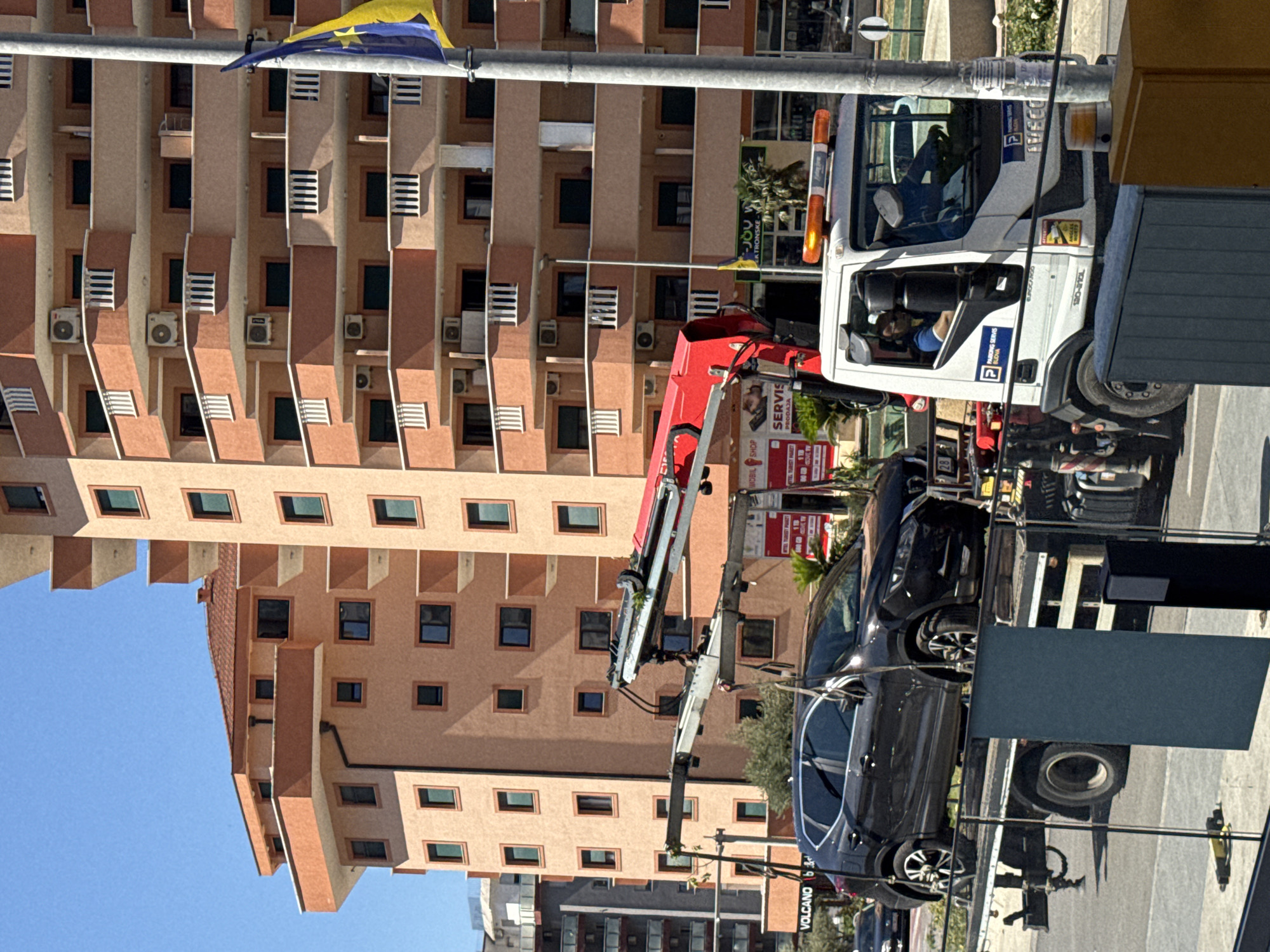 This is a vertically oriented photograph taken in Budva, Montenegro. The scene features a multi-story building with a beige facade, characterized by numerous square windows and balconies, some of which have air conditioning units. In the foreground, a white tow truck with an orange emergency light on top is parked. The truck has branding that includes "Iveco" and "PARKING SERVIS BUDVA". A red tow arm extends from the back of the truck. A black car is partially visible, seemingly being towed or loaded onto the tow truck. There is also a blue and yellow flag visible in the upper left portion of the image. The sky is a clear, bright blue, suggesting a sunny day. The overall impression is of a daytime scene in a commercial or residential area, with a vehicle being attended to by a tow service. Text visible includes "VOLCANO", "MOBIL SHOP", "SERVIS", "EKTRONSKE", "BUDVA", and "PARKING SERVIS".