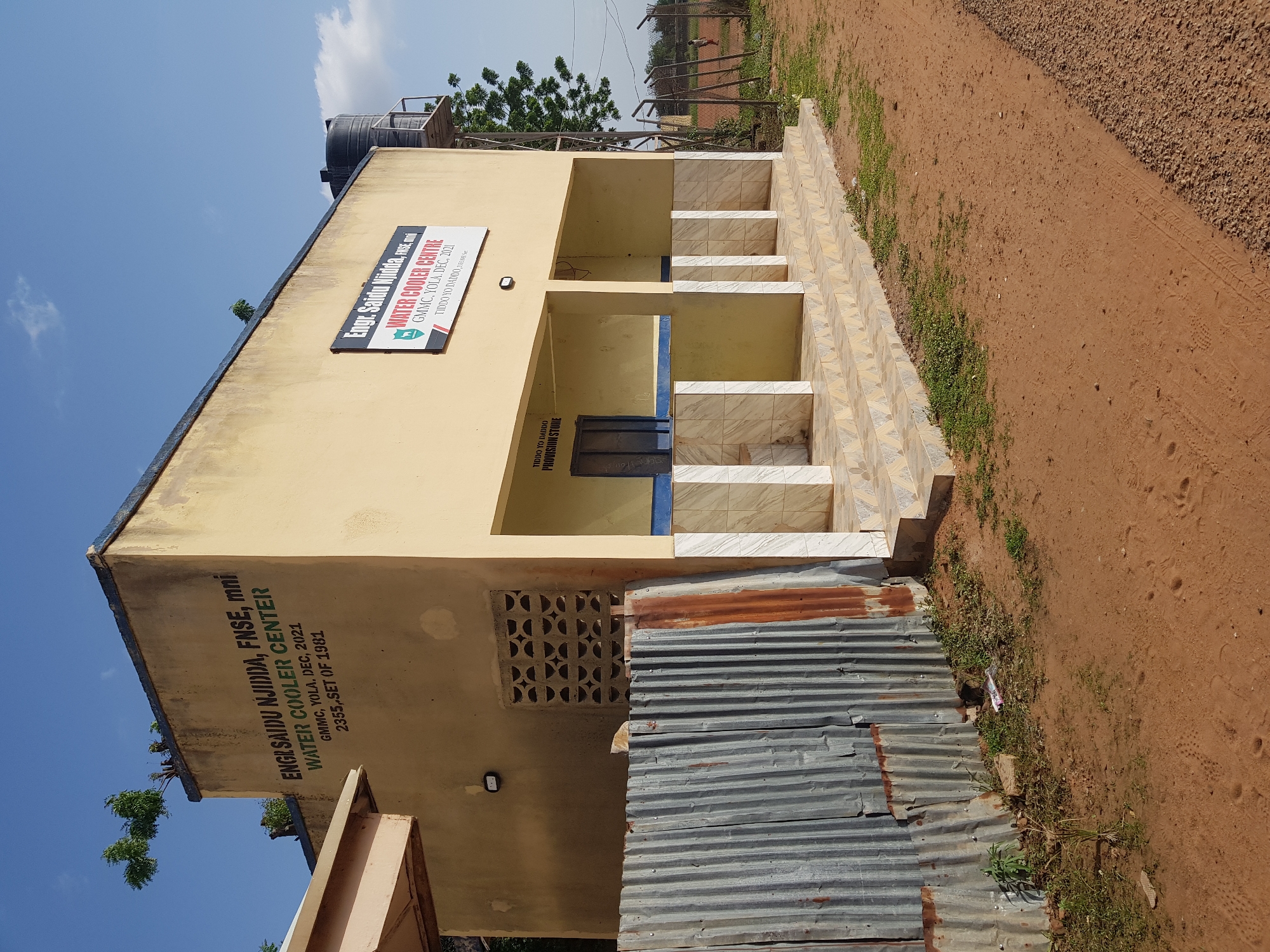 This is a rotated outdoor photograph of a water cooler center building located in Jimeta, Nigeria. The building is light yellow with a concrete structure. A large black water tank sits on a raised platform above the building. The front of the building features several open cubicles with tiled interiors, suggesting they are for dispensing water. A metal grille vent is visible on the side of the building. In front of the building, corrugated metal sheeting has been used to construct a makeshift lean-to.
Two signs are visible on the building. One is a rectangular sign with a logo and text that reads "WATER COOLER CENTRE GMMC, YOLA, DEC, 2021 TIDDO YO DADDO, S". Below this, another sign, partially obscured, appears to have similar text and an emblem. A larger sign on the side of the building also displays text, including "ENGISAIDU NJIDDA, FNSE, mni WATER COOLER CENTER GMMC, YOLA, DEC, 2021 2355, SET OF 1981".
The ground around the building is a mix of reddish-brown soil and gravel, with some sparse green vegetation growing along the edges. The sky is bright blue with a few white clouds, suggesting it is daytime. The overall scene appears to be a functional, perhaps community-oriented facility in a somewhat rural or semi-urban setting.