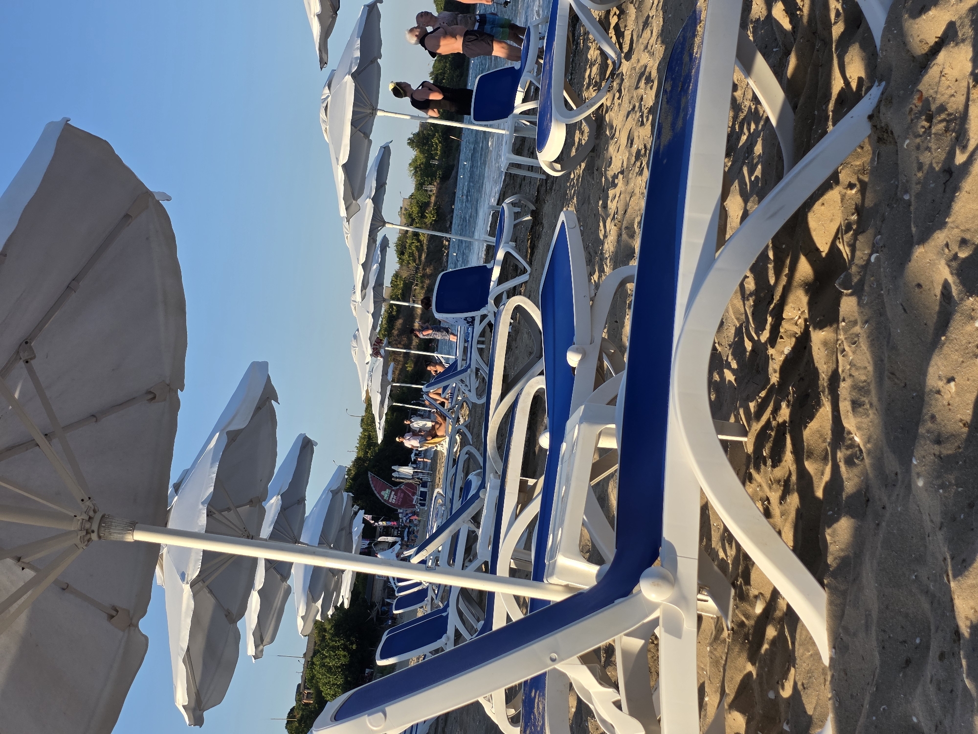 The image depicts a beach scene with several white and blue lounge chairs and white umbrellas set up on the sand. The sea is visible in the background, with small waves breaking near the shore. There are several people on the beach, some standing near the water and others further back. The sky is clear and blue, suggesting it is a sunny day. The location is identified as Chernomorets, Bulgaria. The lighting suggests it is either early morning or late afternoon, with long shadows cast on the sand. The overall impression is a peaceful and inviting beach setting.