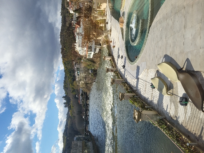The image depicts a riverfront scene in São Pedro do Sul, Portugal, under a partially cloudy sky. The Vouga River occupies the left side of the frame, flowing from the background towards the foreground, with visible rapids and sun reflections on its surface. A multi-arched stone bridge crosses the river in the mid-distance.
On the right side of the image, a paved promenade extends parallel to the river. Three empty, curved chaise lounges are positioned on this promenade: two light-colored and one dark-colored. Adjacent to the promenade, a multi-tiered circular water feature, possibly a thermal pool or fountain, displays turquoise water with an active central jet. A stone balustrade with dark metal railings and green foliage separates the promenade from the river.
Buildings line both banks of the river beyond the bridge. On the left bank, modern structures are visible. On the right bank, a cluster of white and light-colored buildings with multiple stories and balconies extends along the river and up the hillside. The background features densely tree-covered hills under a sky with both blue patches and large cumulus clouds. No individuals are present in the scene.