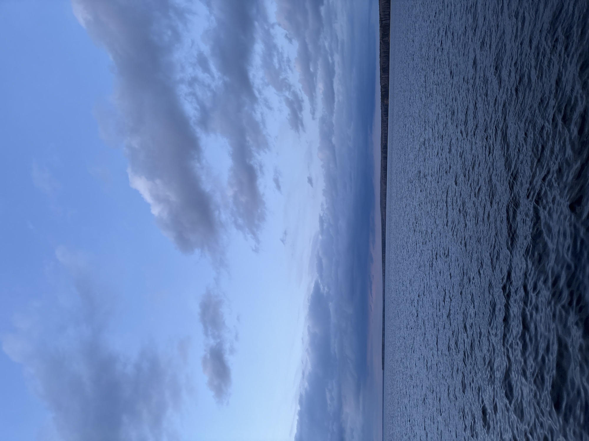 The image depicts a body of water, likely a lake or reservoir, adjacent to a sky with clouds. The sky exhibits a gradient of blue, with some areas showing darker, stormier clouds. A thin line of trees can be seen in the distance. The water surface is ruffled by wind or current. Given the prompt, this is likely near the city of Chaikovsky, Russia.