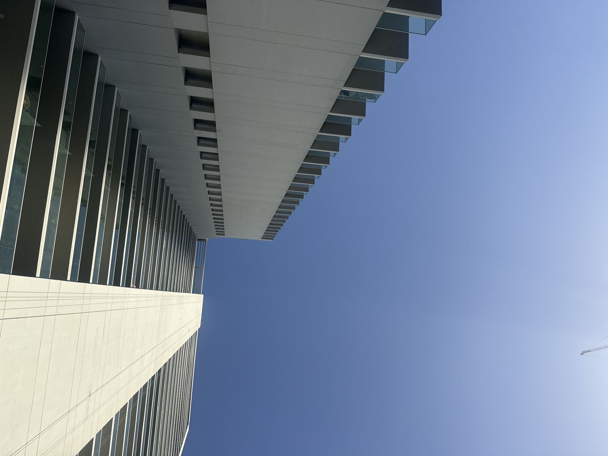 The image captures an upward view of a modern architectural building in Business Bay, United Arab Emirates. The building features a sleek design with linear patterns created by windows and balconies. The facade has a predominantly white and gray color scheme. The sky is clear and blue. A crane is visible in the far background.